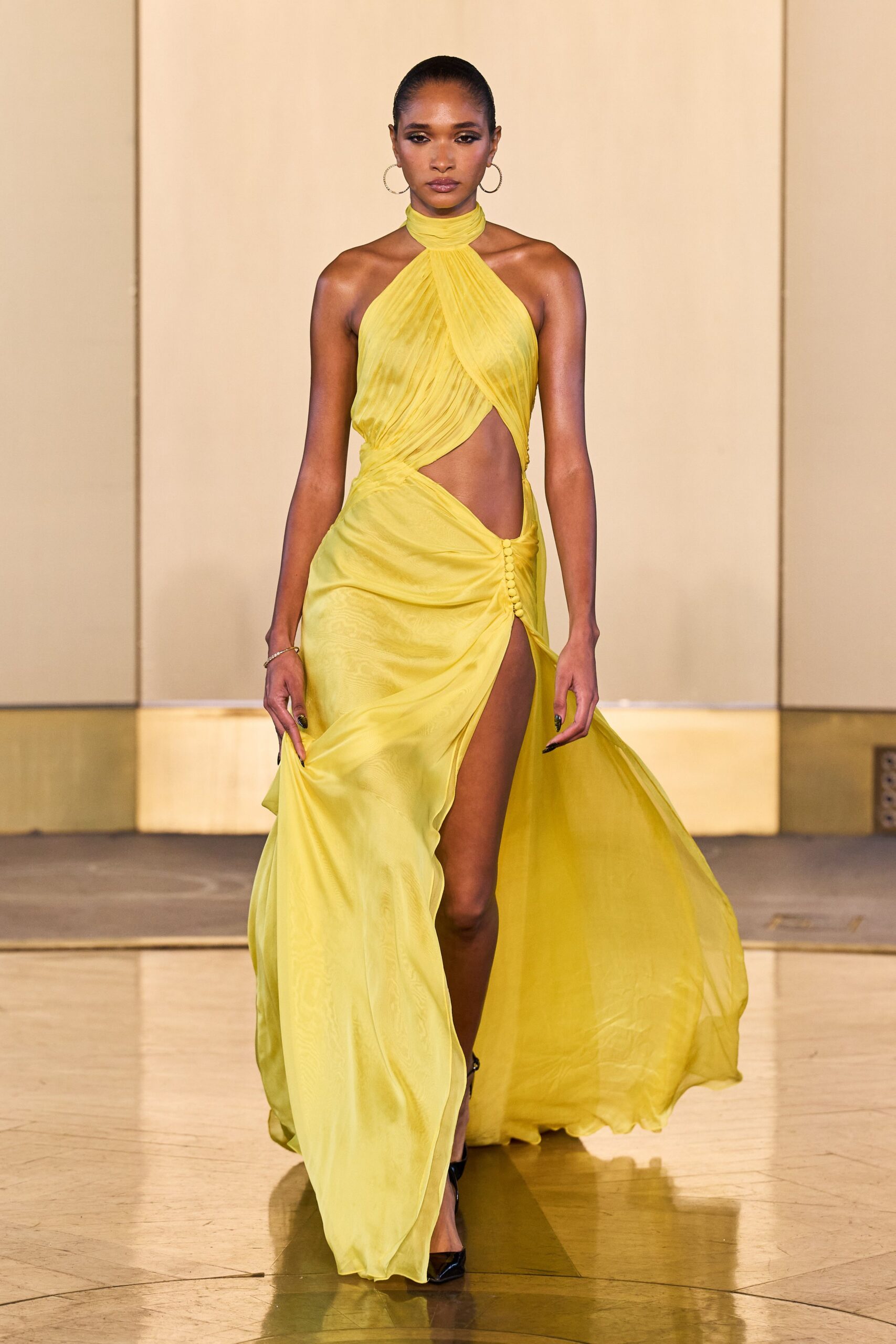 Lemon chiffon halter gown, finely pleated and crossed over a bare waist. Covered buttons climbing the hip beside a thigh-high slit. Thin silver hoops. Black patent pointed pumps. Hotel ballroom air-con after midnight, hem still moving from the stairs.