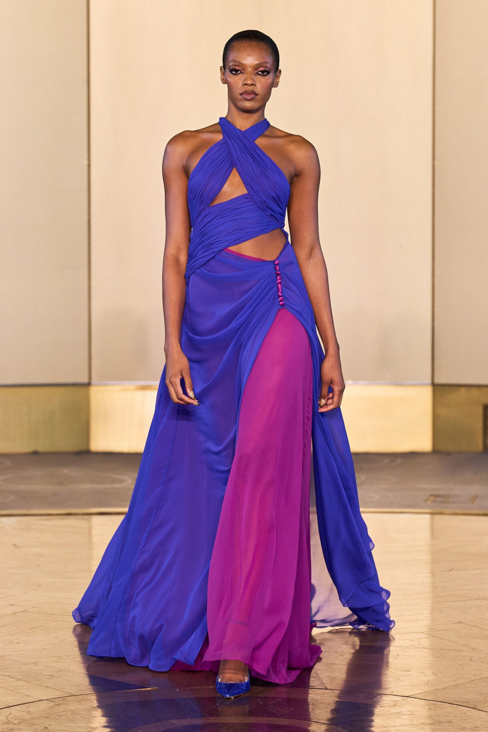 Cobalt chiffon gown cross-wrapped at the neck, cutouts and fuchsia underlayer flashing through. Tiny satin-covered buttons climbing the draped hip split. Blue glitter platforms peeking from the pooled hem. For arriving at a hotel wedding after the ceremony, when the band has started.