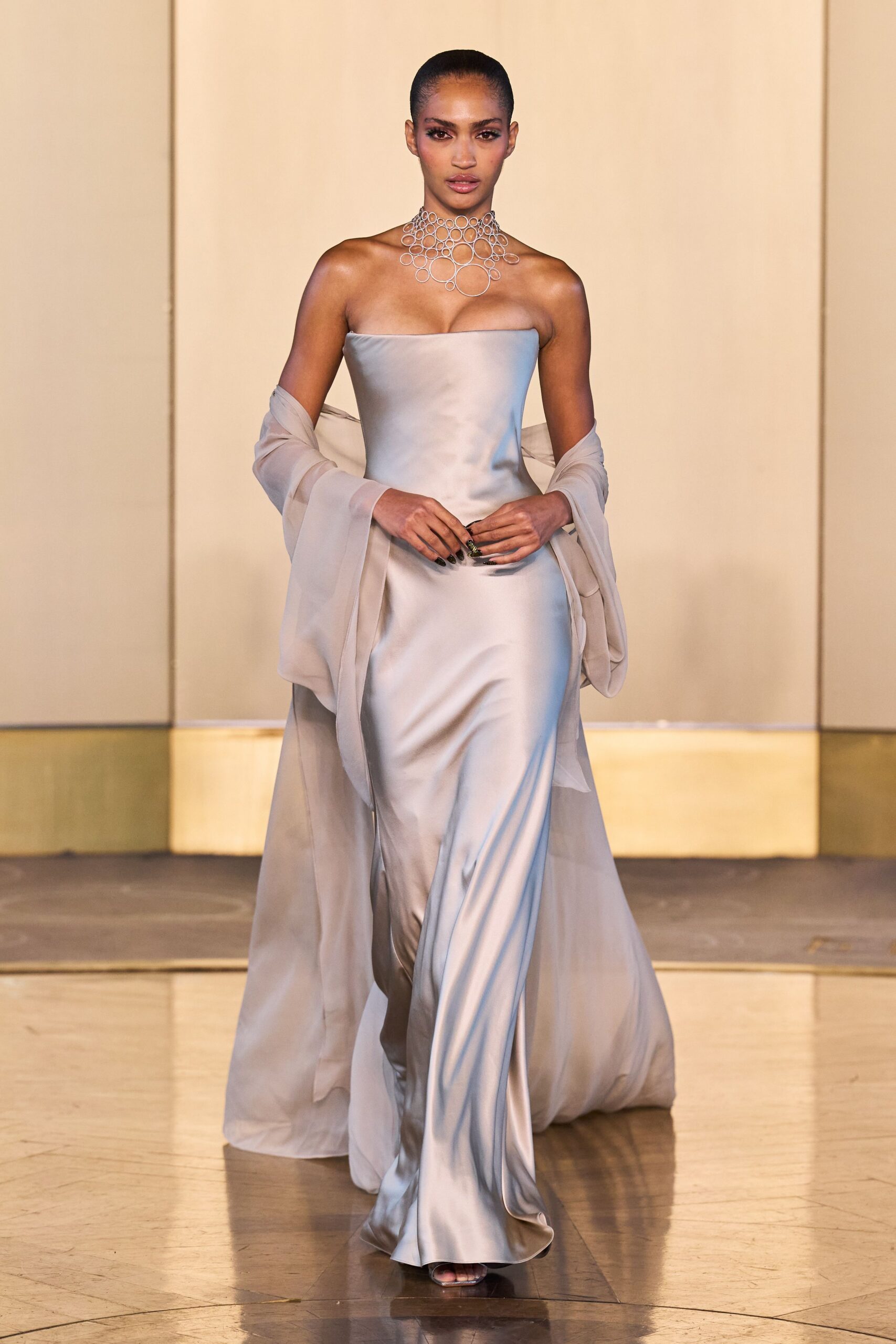 Silver ring-mesh choker cascading in uneven open circles. Pale dove satin column gown, strapless neckline cut straight across the bust. Sheer chiffon stole slipping low off both arms. Barely there metallic sandals peeking under the pooled hem. For crossing a hotel ballroom after midnight with your shoes already in hand.