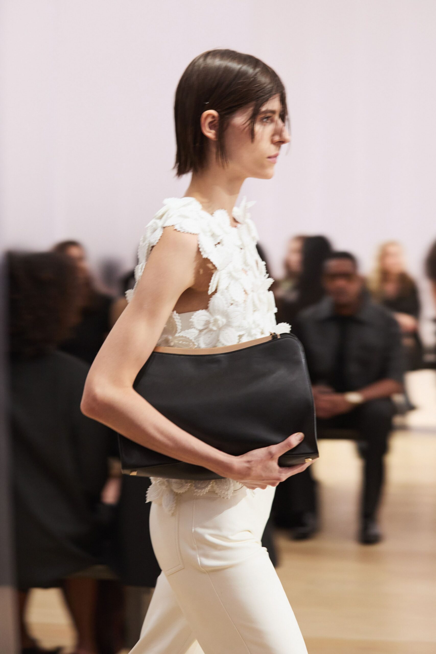 Ivory sleeveless top buried in raised floral lace, petals lifting off the shoulder. Oversized black leather portfolio clutch with a pale binding edge. Cream slim trousers with pressed seams and a single welt pocket. Crossing a museum atrium before the donor lunch starts.