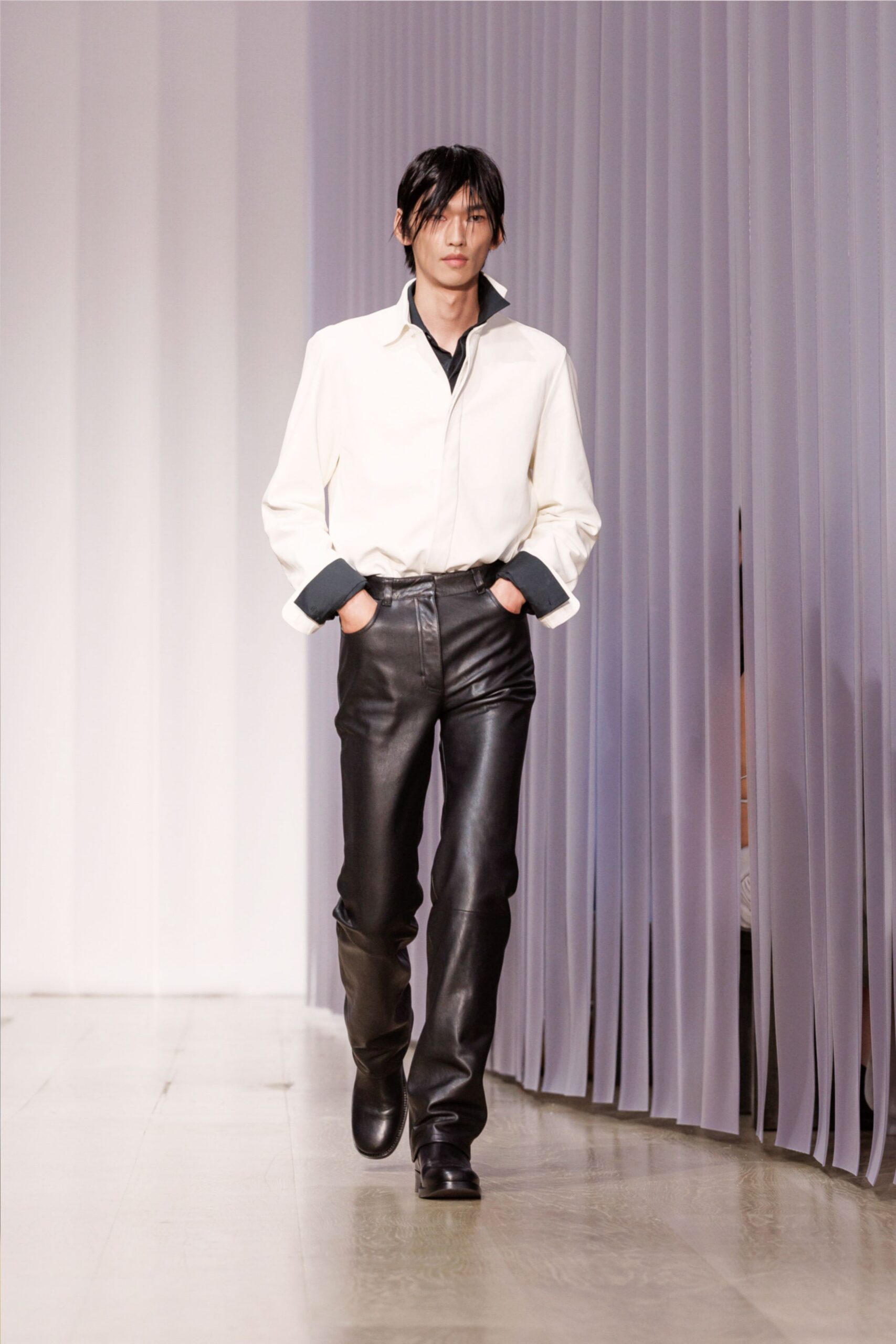 Ivory shirt with a concealed placket and charcoal inner collar turned up. Black leather trousers, pressed close through the thigh with a hooked waistband. Square-toe black boots disappearing under the hem. Walking into a hotel lobby before breakfast service has started.