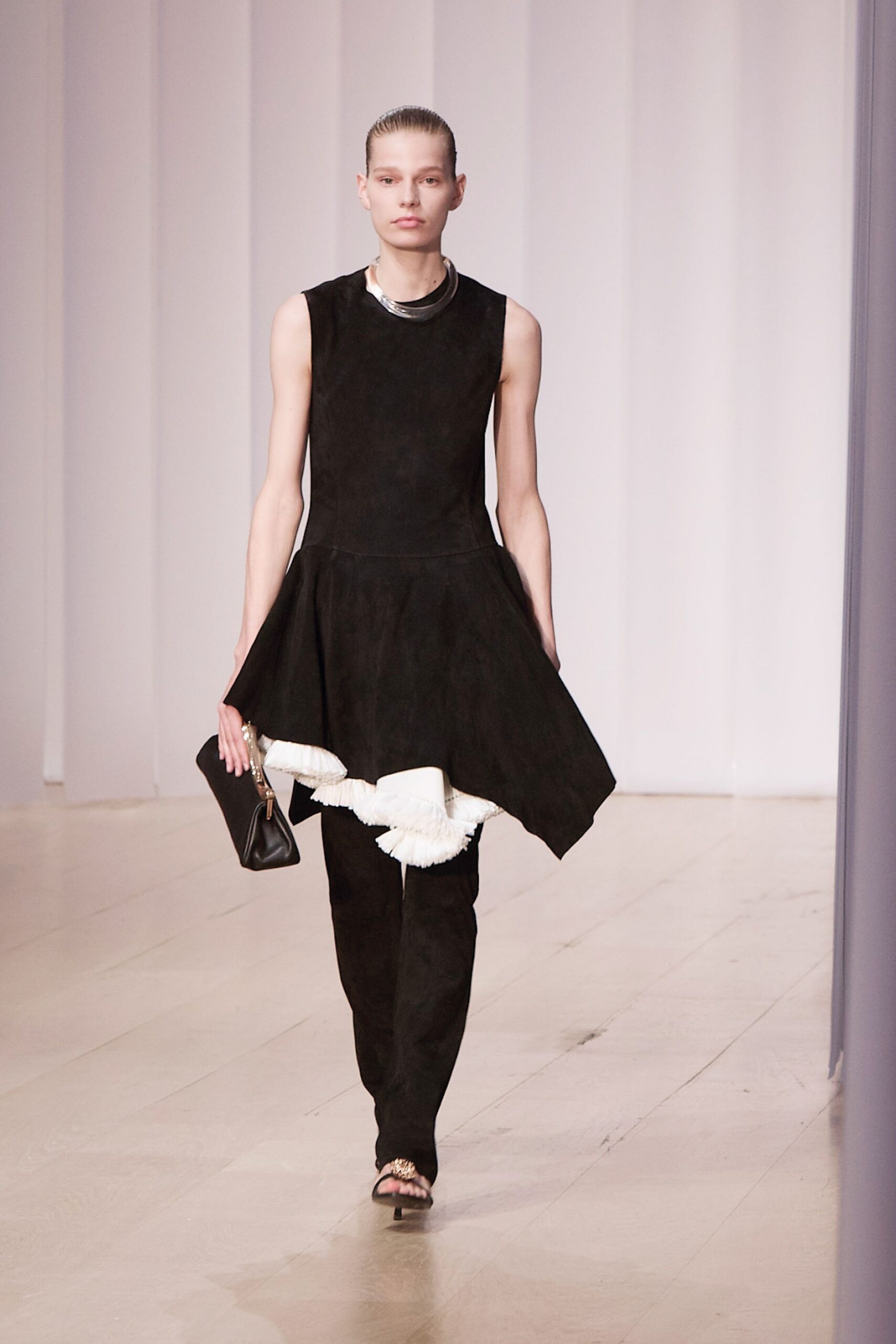 Thick silver collar, doubled at the front. Black sleeveless drop-waist dress with a handkerchief peplum over white fringe. Slim black trousers under the uneven hem. Small black pouch with a chunky chain handle. Open-toe black mules, gold cluster at the vamp. For crossing a museum atrium while the florist is still sweeping up.