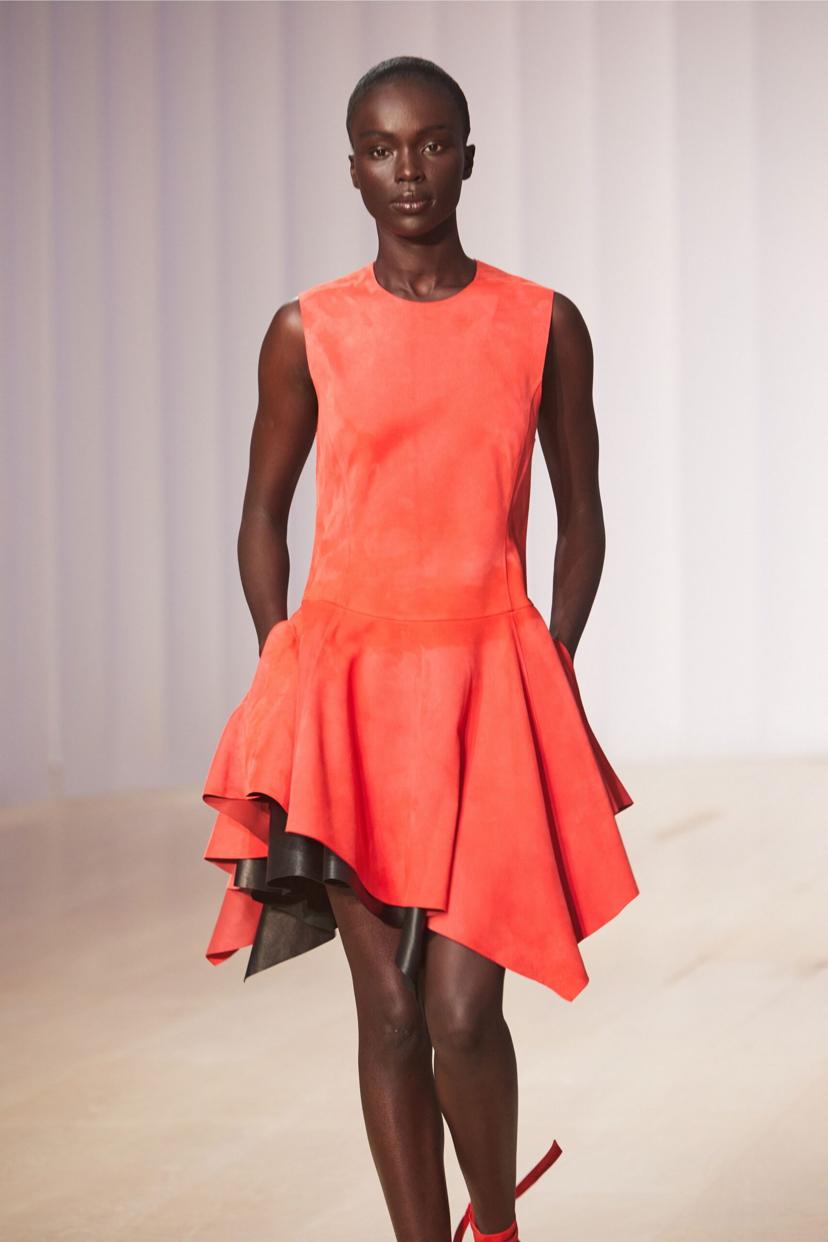 Watermelon satin mini dress with a dropped waist and black-backed flounce. Red ankle-wrap heels, one ribbon tail skimming the floor. Walking across a hotel atrium before the flower delivery arrives.