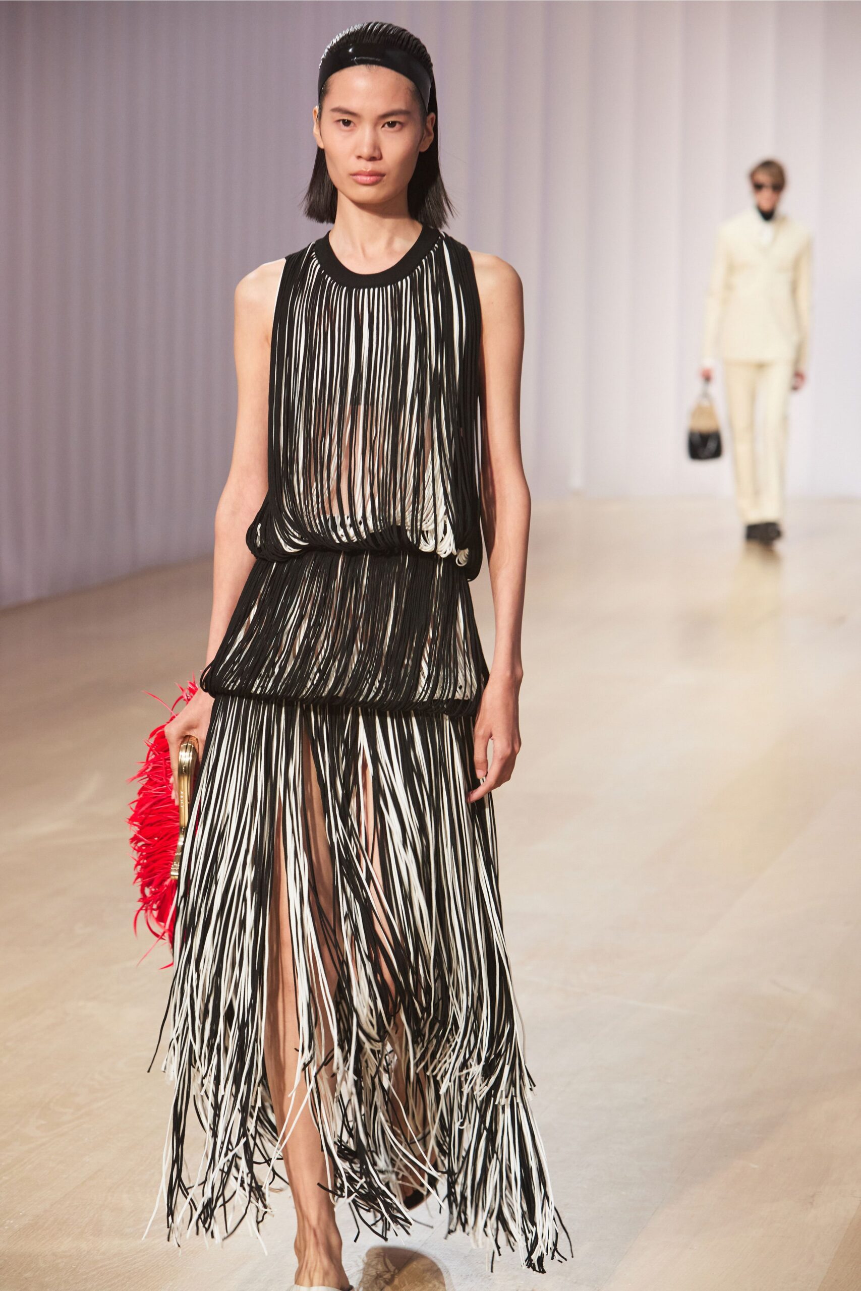Glossy black ridged headband holding the hair flat. Sleeveless column built from black and white cords, pooled in tiers at the waist. Red feathered clutch with a curved gold frame. Pointed cream mules peeking through the swinging hem. Crossing a hotel atrium while the pianist is still tuning up.
