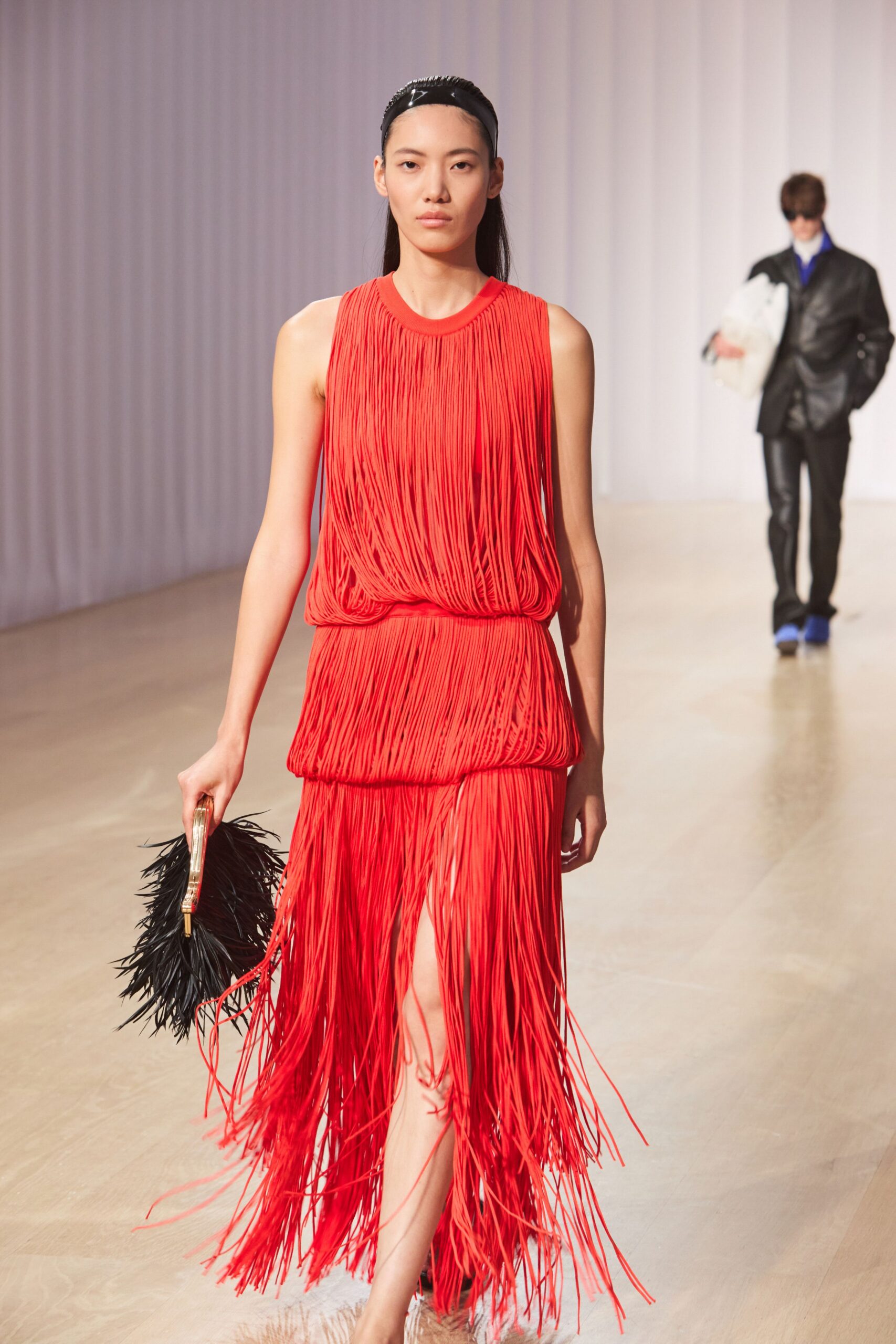 Glossy black patent headband, narrow ridges catching the light. Red sleeveless dress built from dense cord fringe, blousing at two dropped seams. Black feather clutch with a clear lucite frame and gold clasp. Barely there nude sandals, lost under the moving hem. Walking into a hotel ballroom just after the band has started.