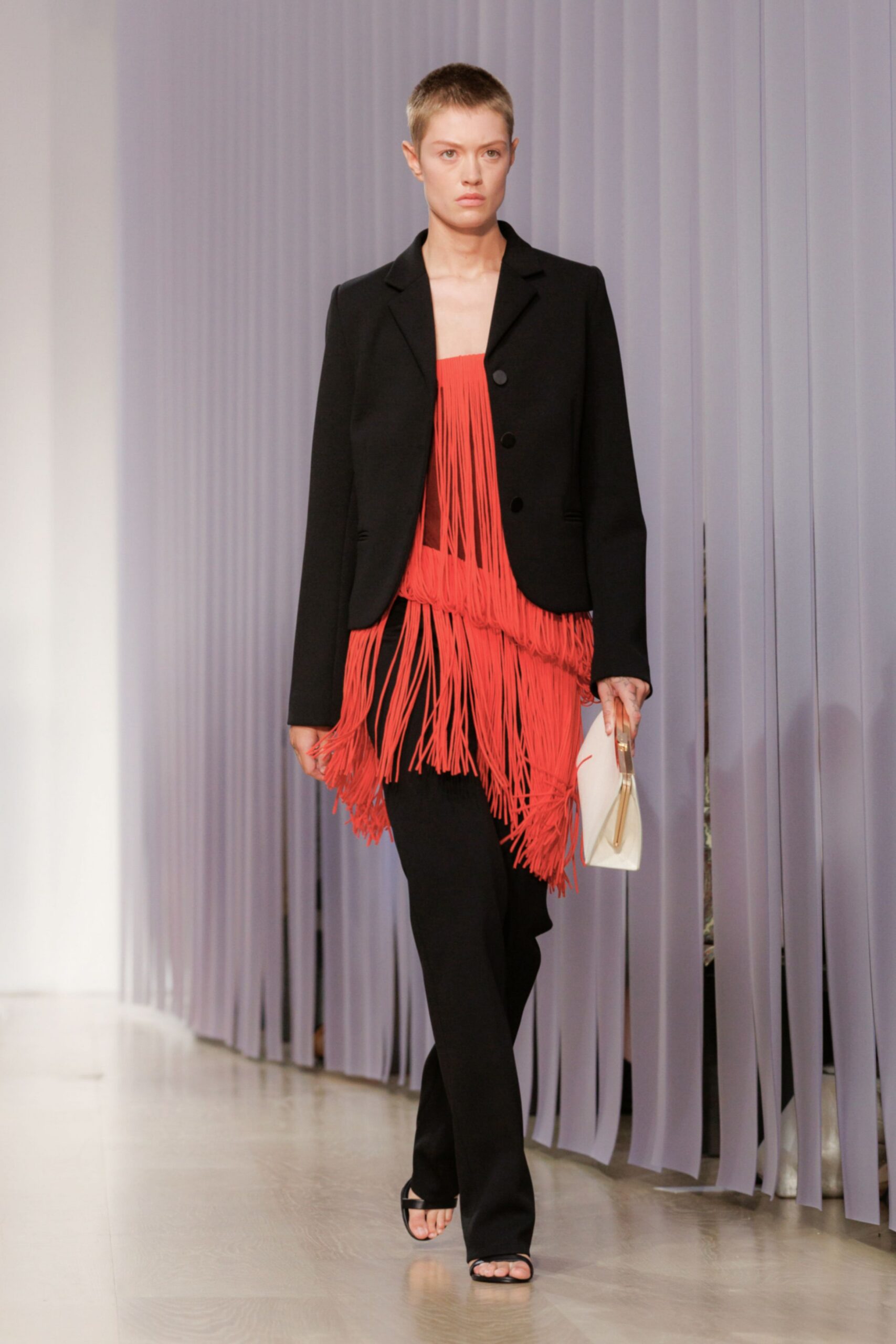 Black tailored blazer with three matte buttons and a curved front hem. Coral rope-fringe dress, the strands layered thick across bust and hips. Black straight trousers breaking softly over the instep. Ivory folded clutch with a rigid gold frame. Black thong heeled sandals. Walking into a hotel bar before the pianist has started.