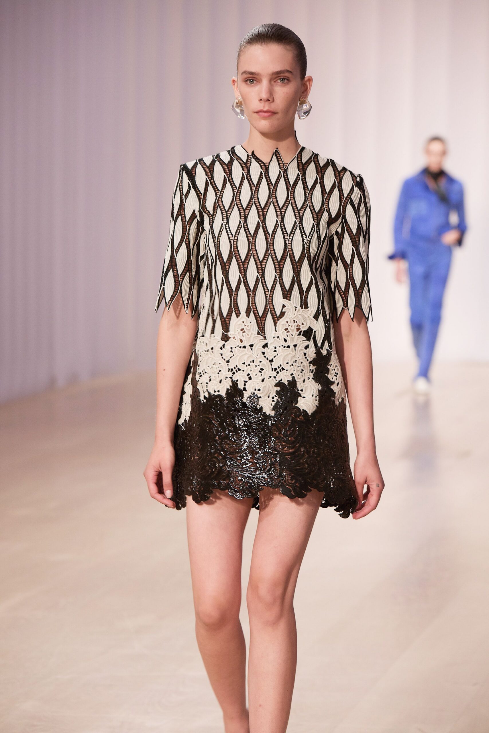 Clear sculpted drop earrings catching light at the jaw. Ivory mini dress over black tulle, flame-shaped openwork and scalloped guipure hem. Walking into a hotel atrium while the florist is still wiring the centerpieces.