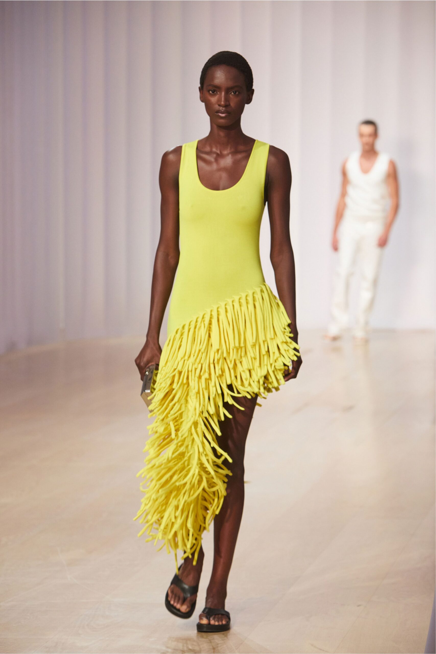 Acid-yellow sleeveless knit dress, one side exploding into long tubular fringe. Small box clutch with mirrored sides. Black thong sandals on a low flatform sole. Crossing a hotel terrace at noon with the pool still closed.