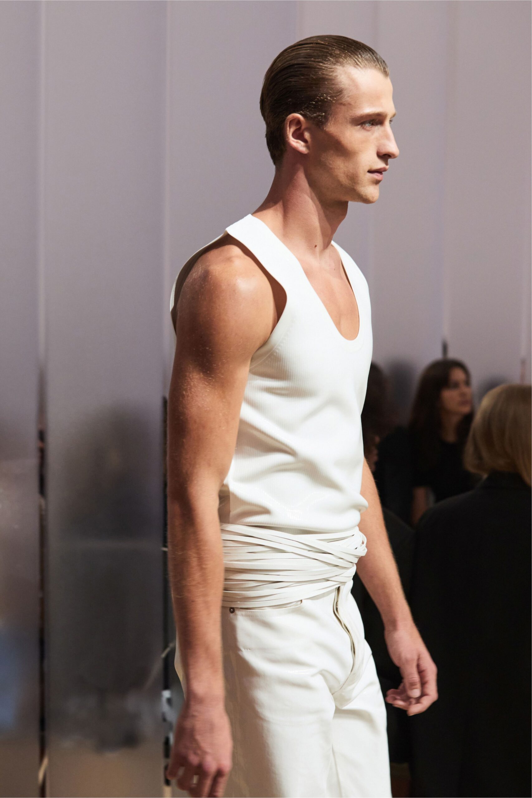 White ribbed tank with a scooped neck cut wide at the shoulder. White leather cords wound low around the hips, one end hanging. White trousers with a curved fly seam and a tiny rivet at the pocket. Walking through a sunstruck atrium after swimming somewhere expensive.