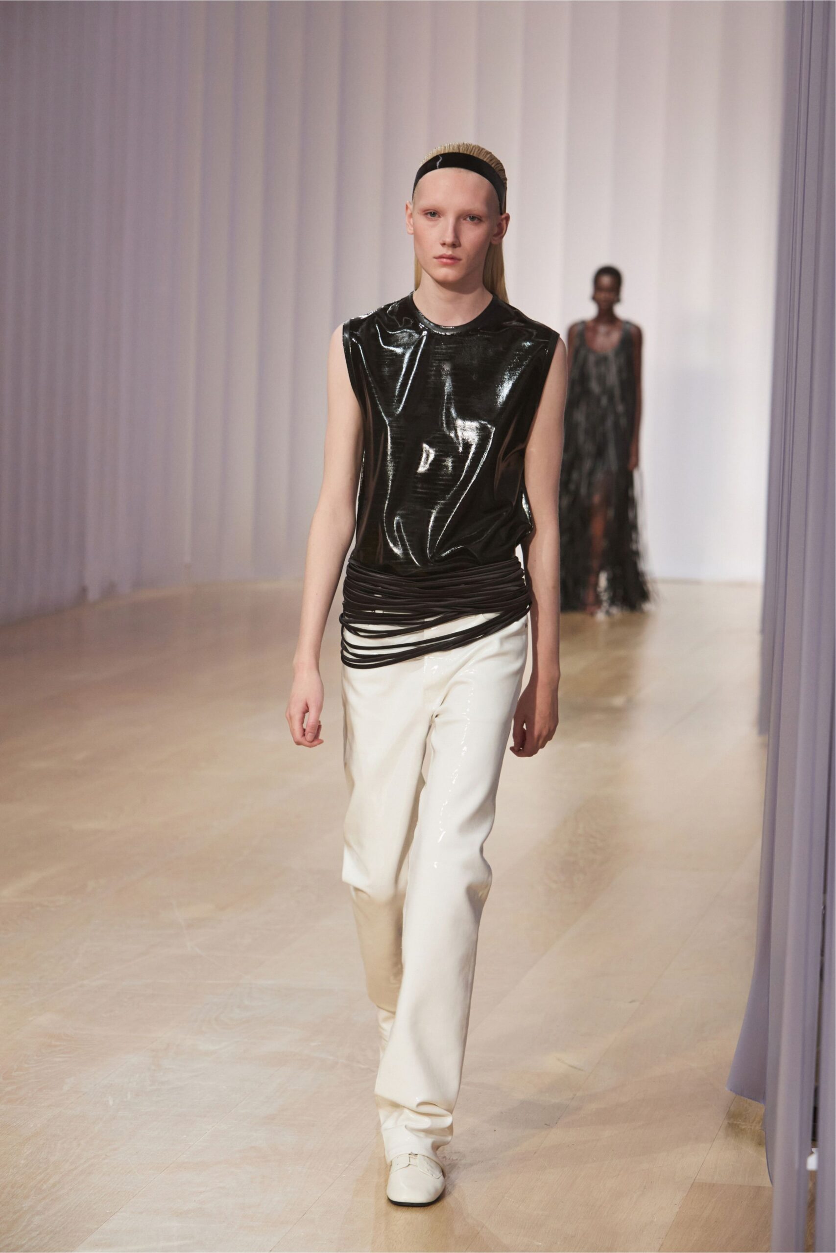 Wide black elastic headband holding the slick hair flat. Black high-gloss sleeveless shell, creased like wet vinyl. Roped black cords looped low around the hips. White coated trousers falling straight with a slight break. Square-toe white shoes with a thin strap. Walking the long corridor to a hotel spa before it opens.