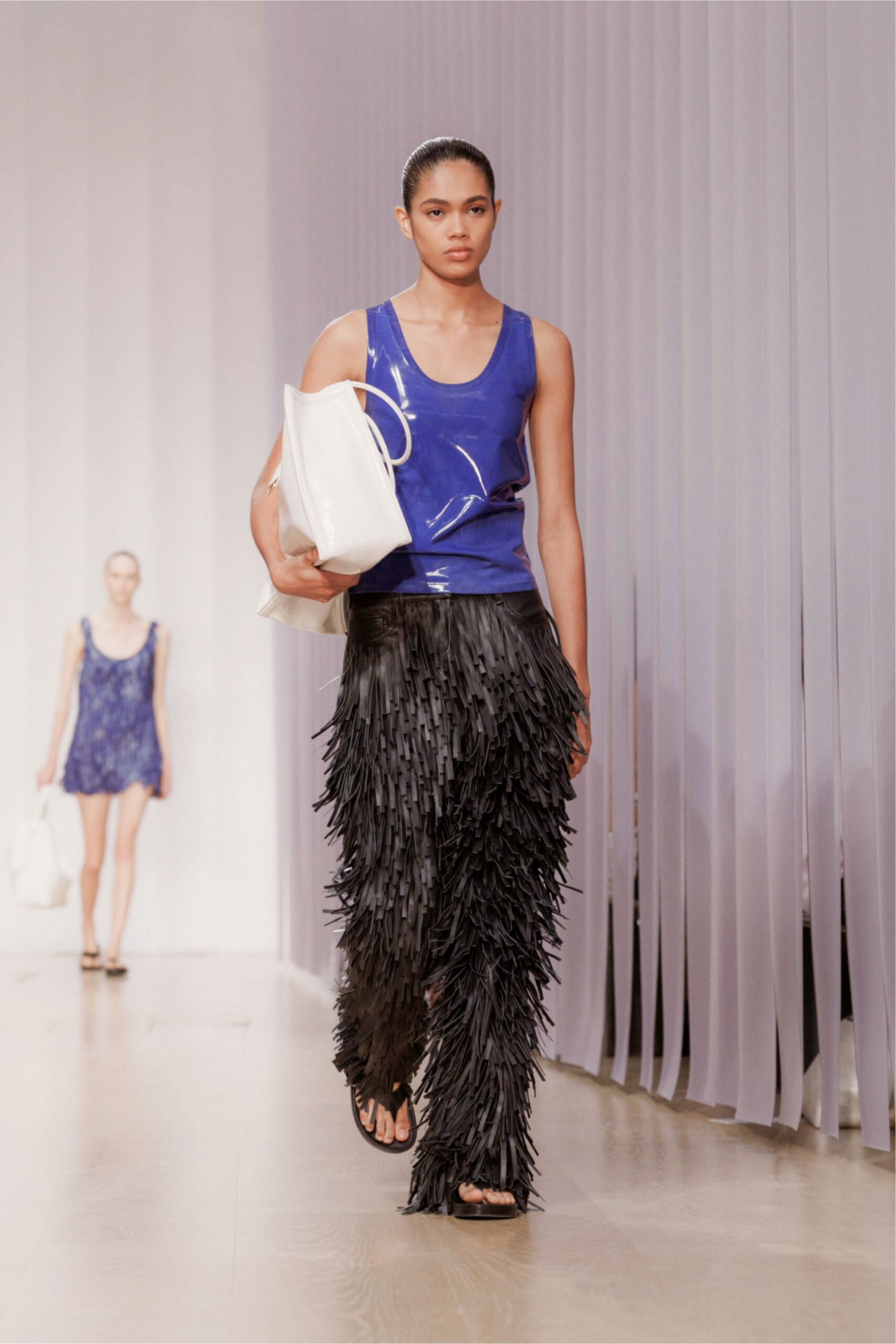 Cobalt coated tank, glossy surface creasing at the waist. Oversized white tote tucked bluntly under one arm. Black trousers buried under dense strip fringe to the ankles. Flat black thong sandals. Walking back from a beach club with wet hair and no towel.