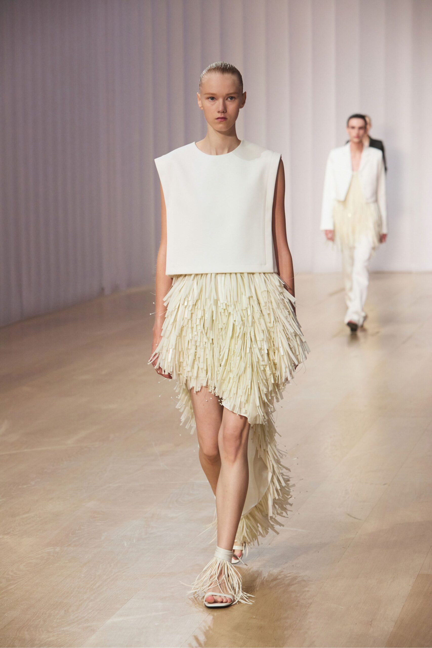 Ivory bonded shell top with squared shoulders and a clean cut edge. Cream fringe skirt, dense strips falling into an uneven trailing hem. Barely there sandals with raffia tassels bursting over the toes. Crossing a sunlit hotel lobby after midnight, carrying your shoes in your head.