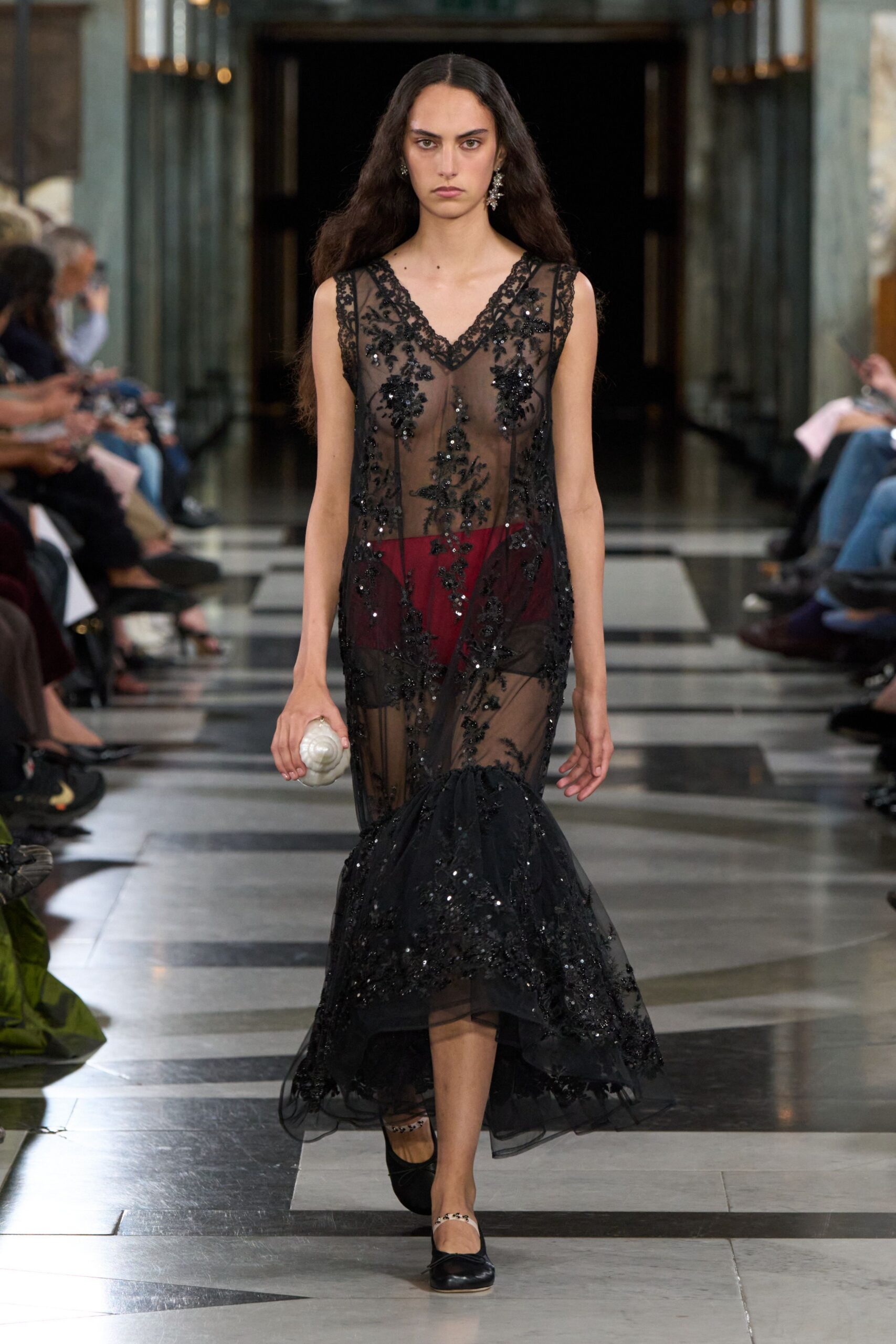 Sheer black tulle gown with beaded floral embroidery and a dropped fishtail hem. Deep garnet briefs showing starkly beneath the mesh. White shell clutch cupped in one hand. Black ballet flats with slim leopard straps across the instep. Crossing a hotel lobby after midnight with salt still on your skin.