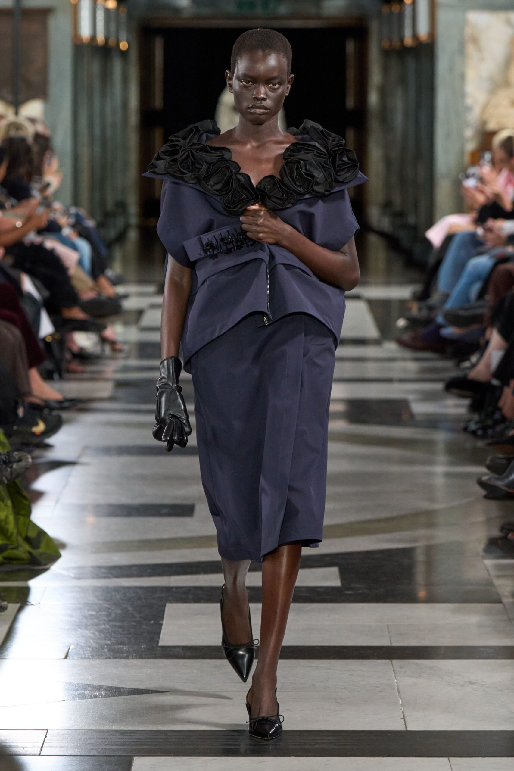 Ink satin top with oversized black rosettes crowding the off-shoulder neckline. Matching wrap skirt, folded panel breaking cleanly across the knees. Single black leather glove. Black pointed mules with tiny bow ties at the toe. Crossing a marble lobby after the vote already turned against you.