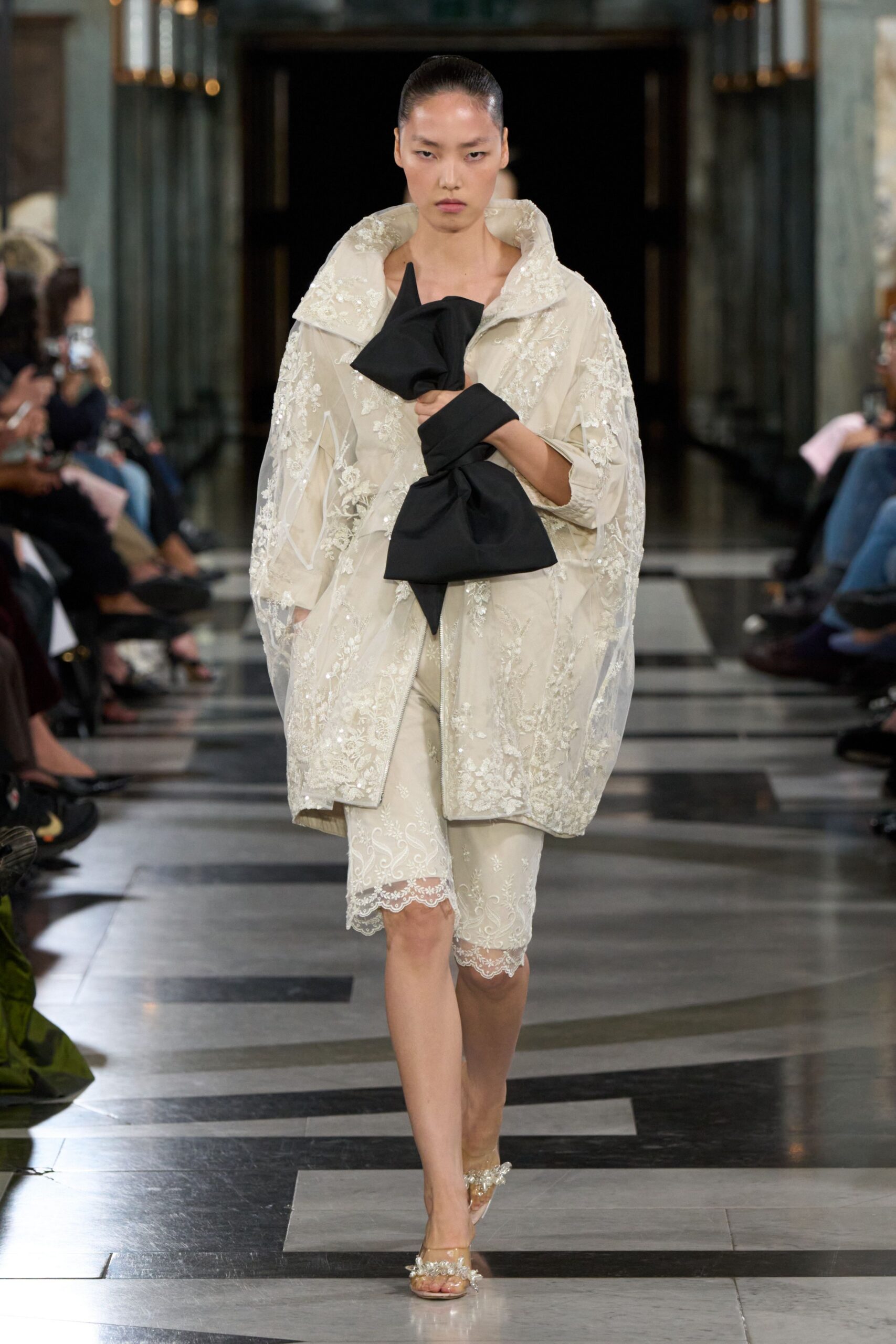 Ivory cocoon coat in sheer tulle, crusted with floral embroidery. Oversized black bow clutch, folded into stiff origami points. Cream lace-trim cycling shorts with a scalloped hem at the knee. Clear heeled sandals topped with clustered crystal blossoms. For crossing a hotel ballroom after the wedding has already thinned out.