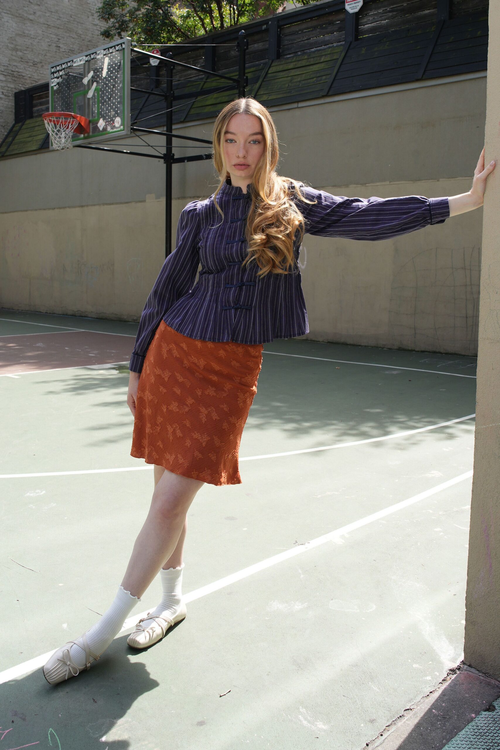 Indigo pinstriped blouse with frog closures and softly puffed sleeves. Rust bias-cut skirt with tonal leafy jacquard and a wavering hem. White ribbed crew socks. Cream ballet flats with a drawstring bow across the square toe. Sunday on the apartment court before anyone starts keeping score.
