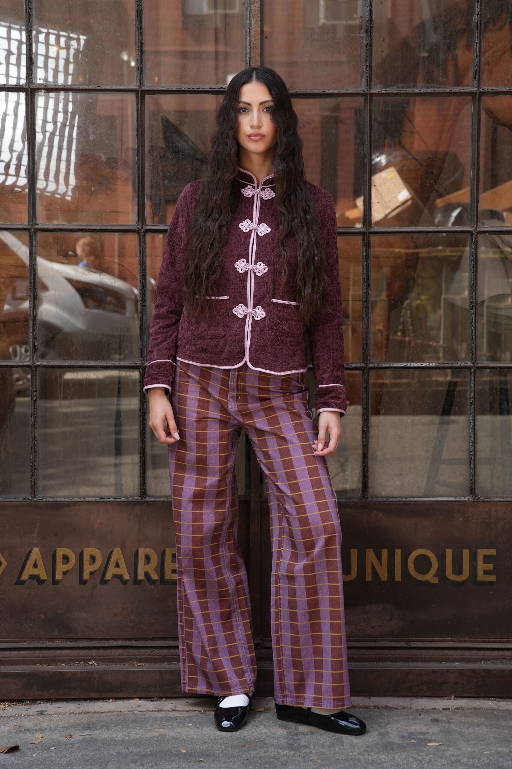 Burgundy boucle jacket with pale frog closures and piped edges. Lilac and brown windowpane trousers, creased straight through the leg. White ankle socks. Black patent flats with squared toes. Outside a Chinatown antique shop before the street has properly woken.