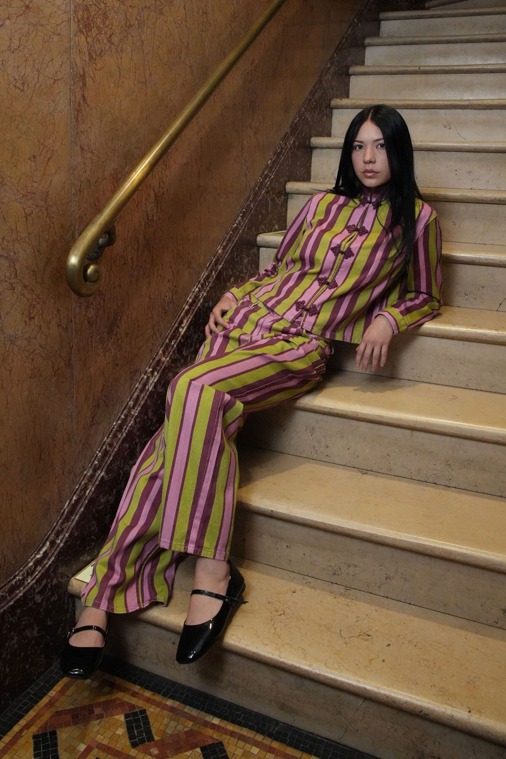 Silky striped jacket in orchid and chartreuse, closed with frog knots. Matching flared trousers, vertical bands breaking around the knee bend. Black patent Mary Janes. Pausing on a marble stair after slipping out of the banquet room.