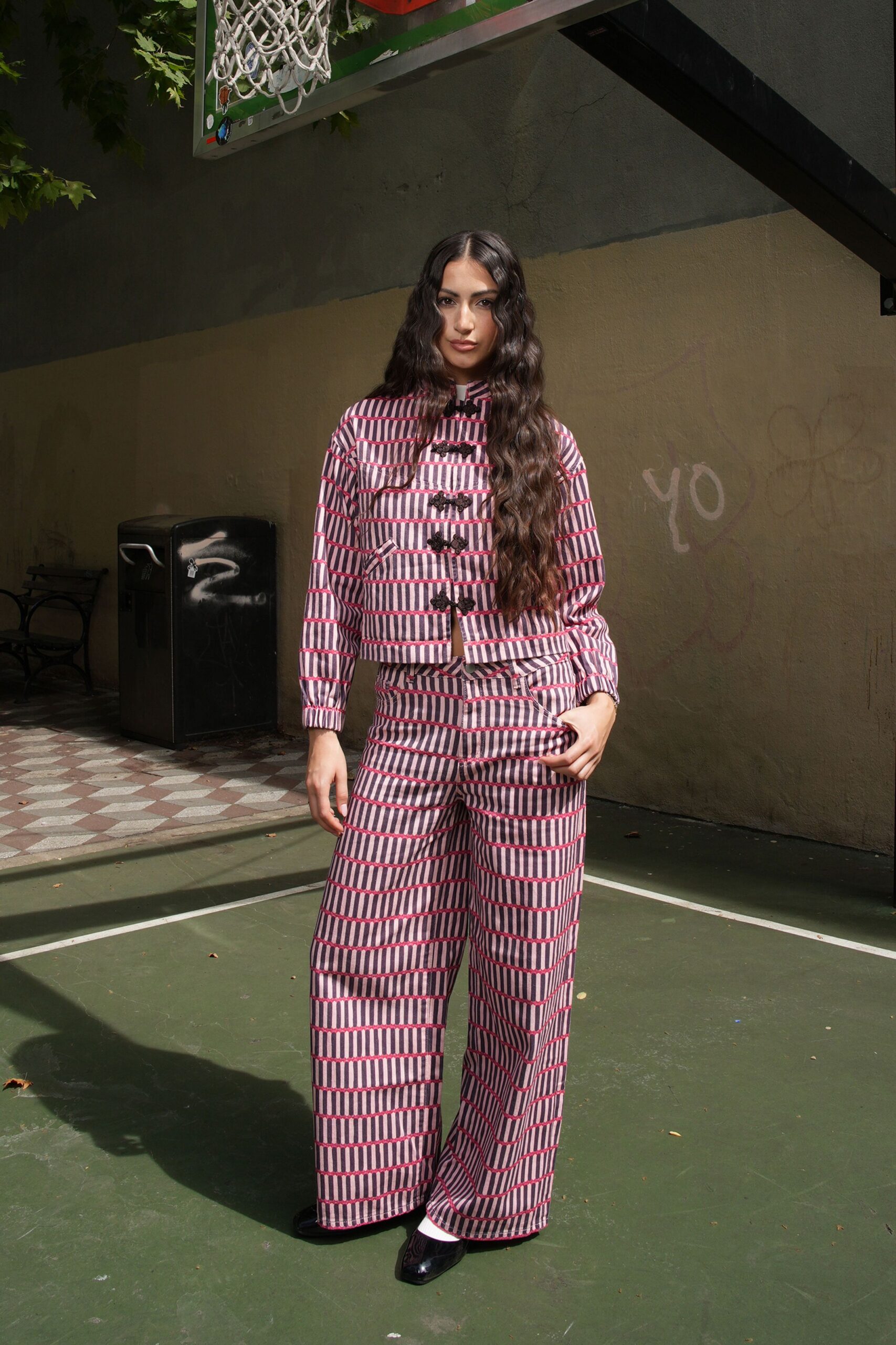 Quilted cropped jacket in pink-and-ink striped checks, frog closures down the chest. Matching wide-leg trousers, creased sharply and pooling over the shoe. Black square-toe flats with white socks flashing above. Afternoon on the neighborhood court after the game has already moved on.