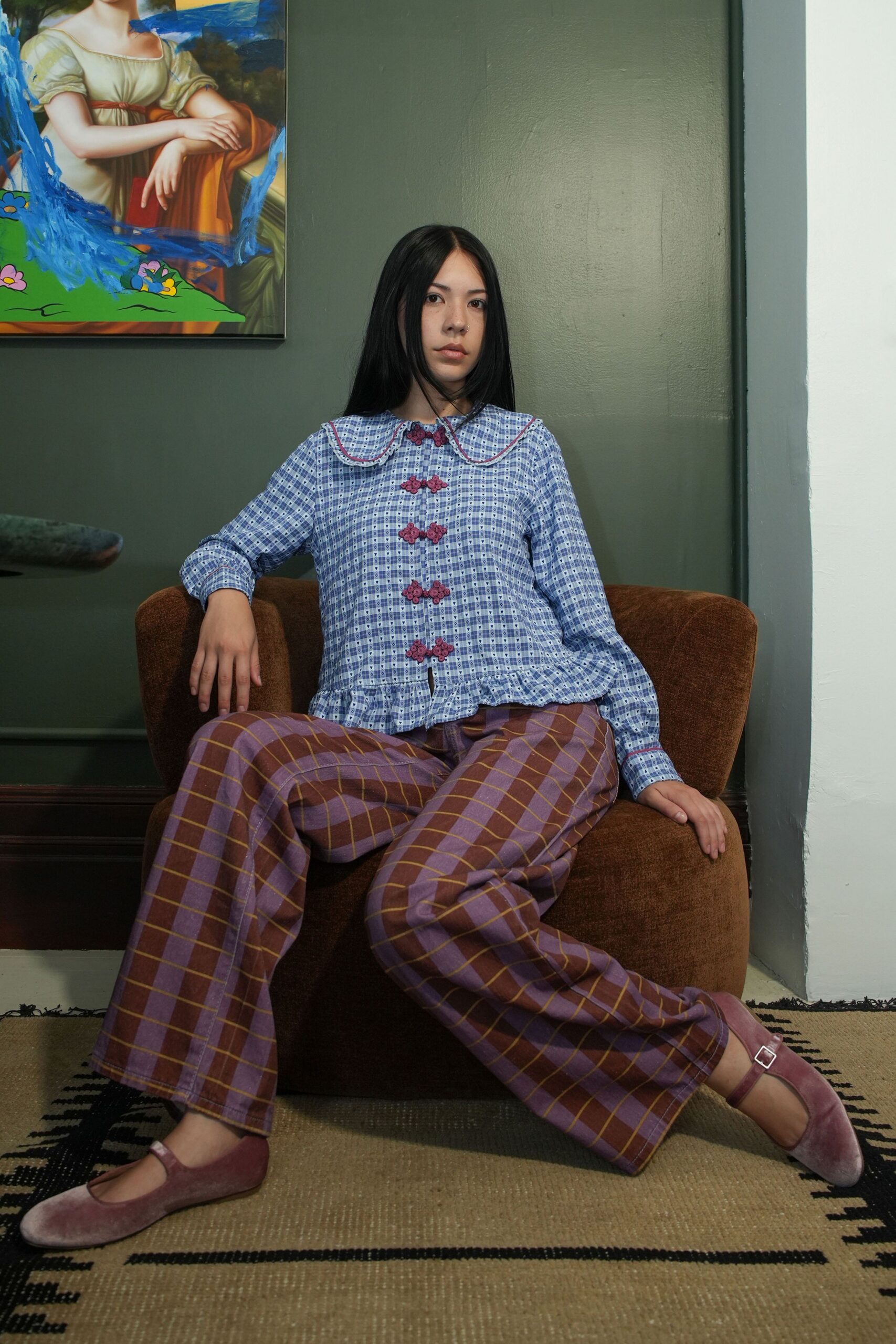 Blue gingham blouse with a scalloped Peter Pan collar and frog closures. Lilac and brown windowpane trousers cut wide through the leg. Dusty mauve velvet Mary Janes with a squared silver buckle. Afternoon in a rented parlour, rain starting against the sash windows.