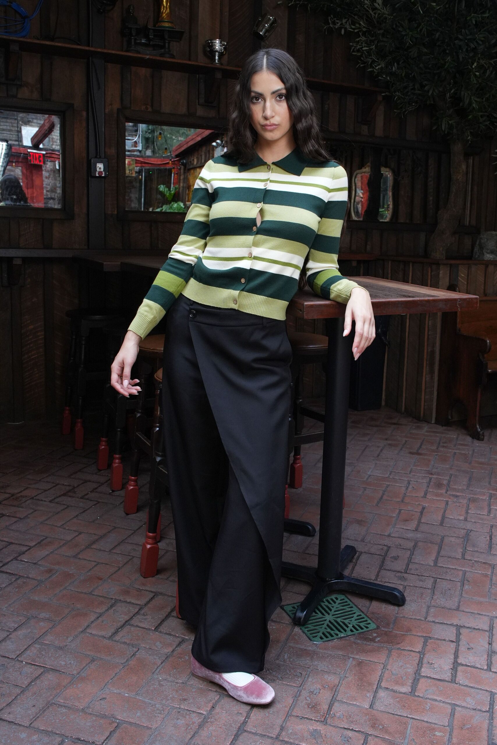 Fine-gauge striped knit cardigan in moss, cream and citron, one button left open. Black wide-leg trousers with a flat front and soft break. Dusty pink velvet flats over white socks. Pub garden at noon, before anyone else has ordered lunch.