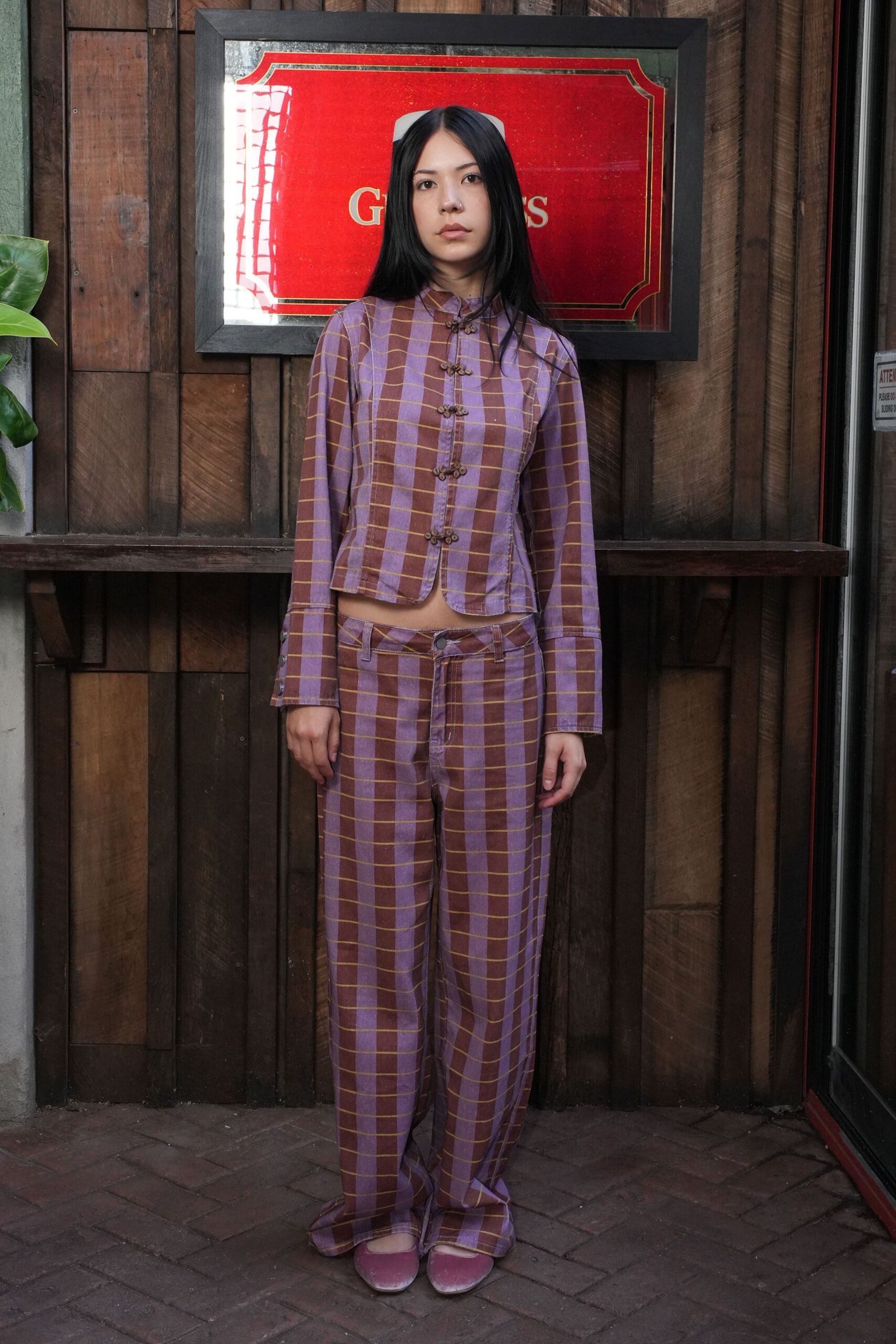 Lilac and cocoa windowpane jacket with frog closures and a slight peplum flare. Matching low-rise trousers, the fly skewed slightly off center. Dusty mauve velvet flats with rounded toes. Outside a private dining room before the first bottle is opened.