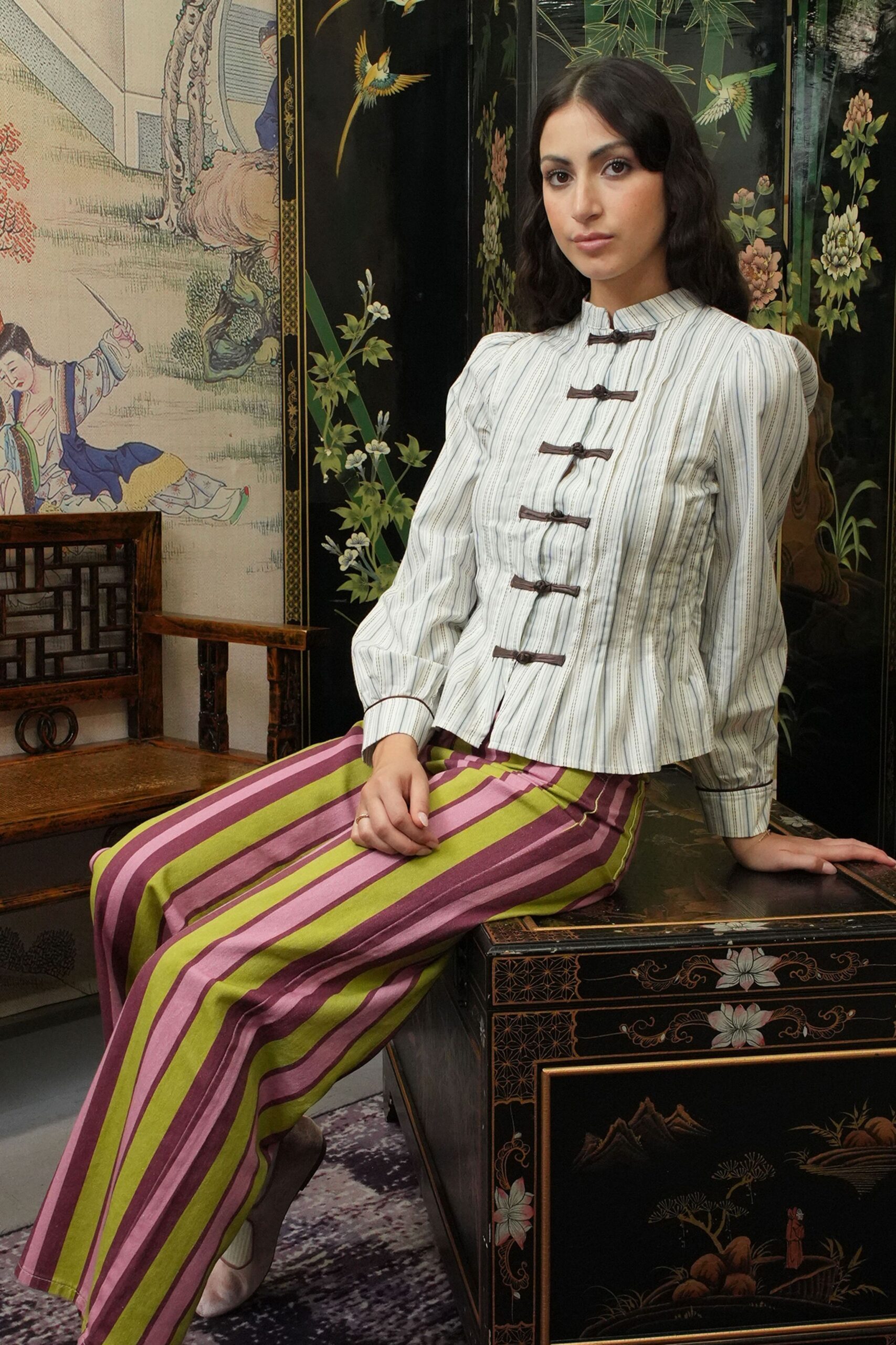 Ivory striped blouse with frog closures and puffed shoulder heads. Wide trousers in lime and mauve satin stripes, crease running sharp. Pale pumps with a rounded vamp. Afternoon in a lacquered sitting room, waiting for tea to be poured.