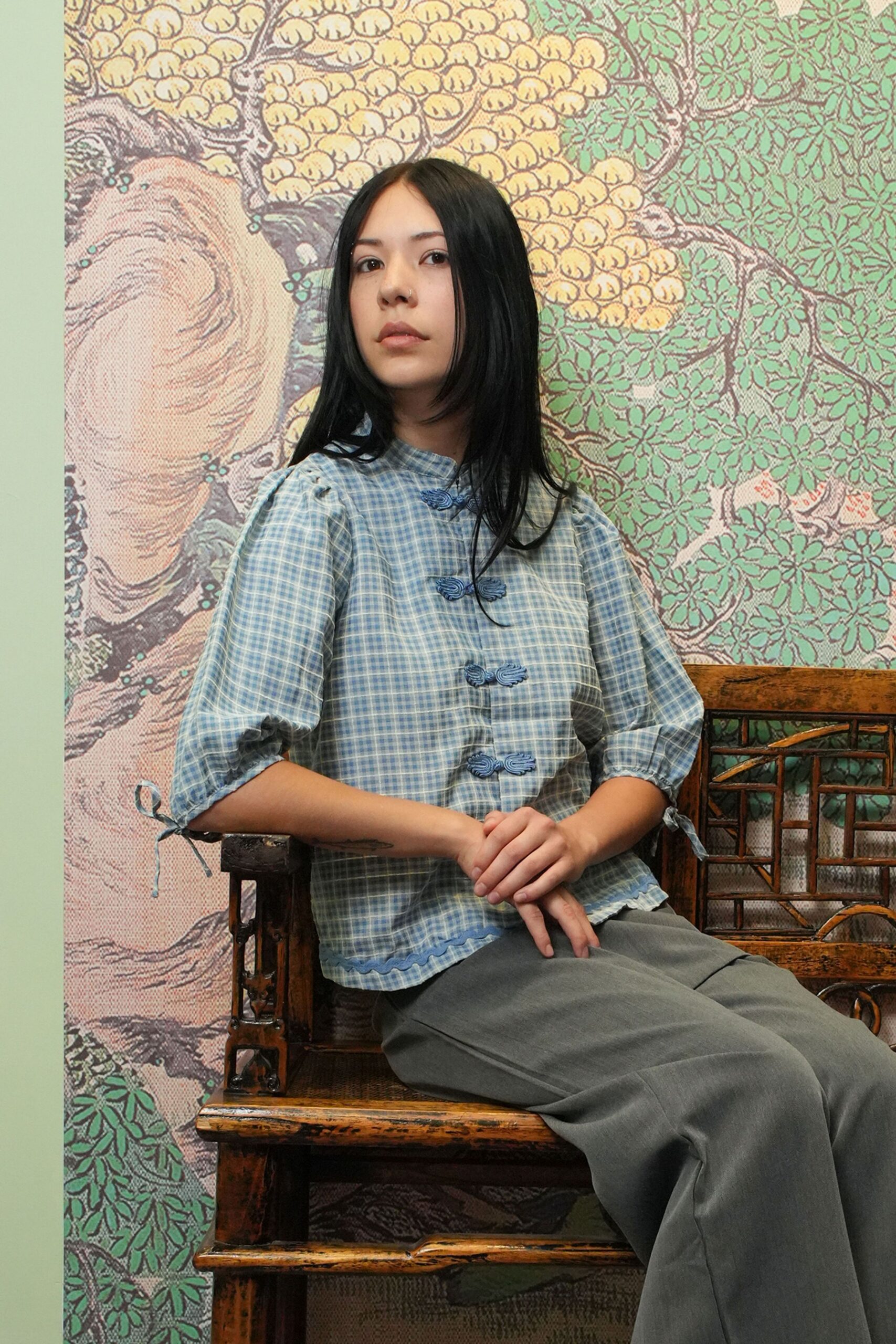 Powder-blue gingham blouse with frog closures and scalloped ric-rac hem. Soft slate trousers falling loose over the lap. Sitting in a tea house under mural wallpaper, waiting out the hottest part of afternoon.