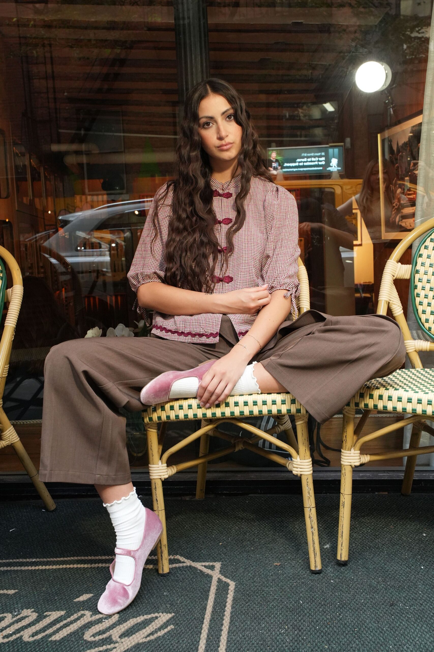 Burgundy micro-gingham blouse with frog closures and scalloped ric-rac hem. Taupe wide-leg trousers breaking softly over the chair edge. White ankle socks with a lettuce-frill cuff. Pink velvet Mary Janes, strap cutting across the instep. Waiting out the drizzle at a cafe table before the matinee starts.