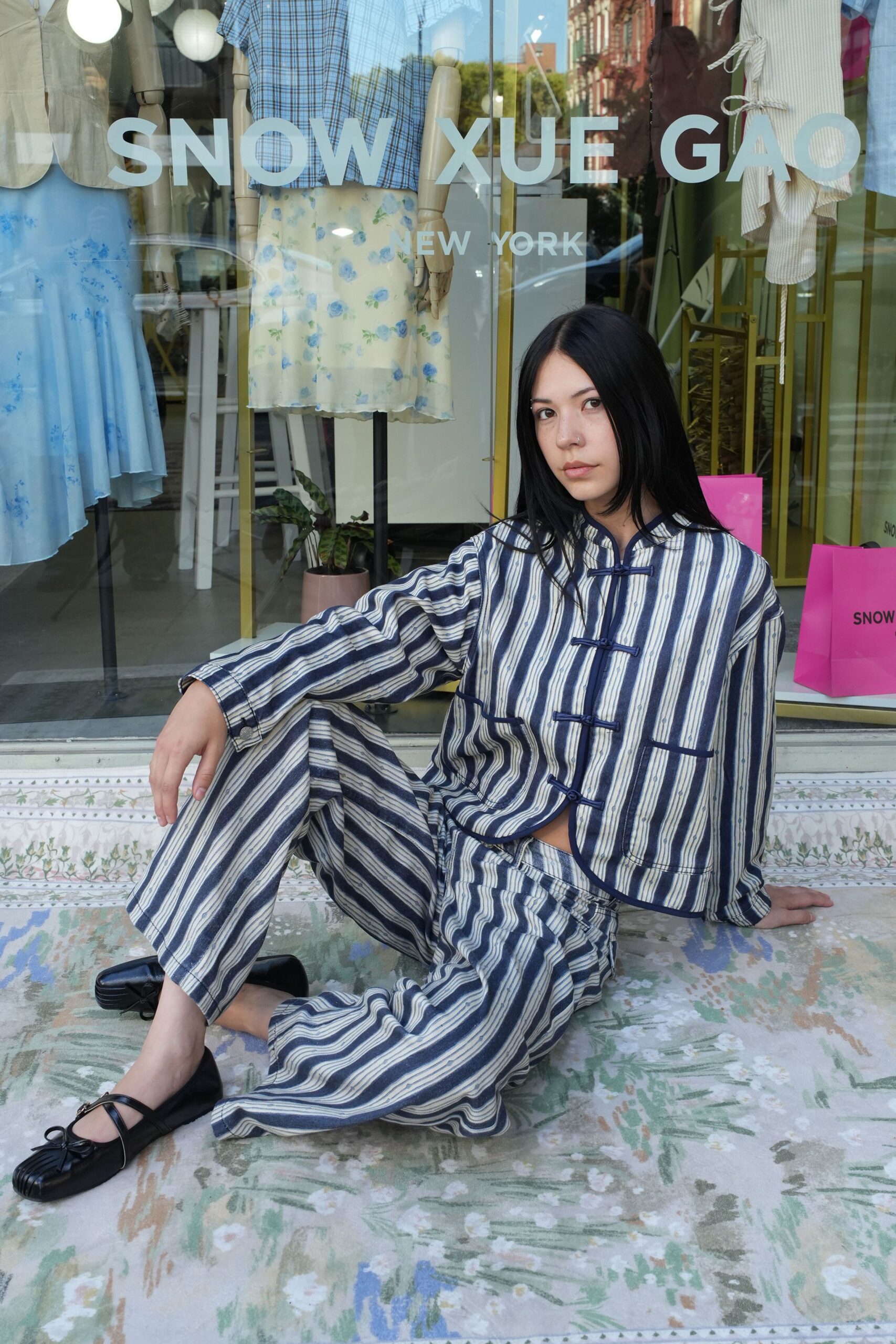 Boxy indigo-and-ivory striped jacket with frog closures and piped edges. Matching wide trousers in the same woven stripe, hem breaking softly. Black square-toe Mary Jane flats with a tiny bow. Sitting on a shop step after buying peaches at the Saturday market.