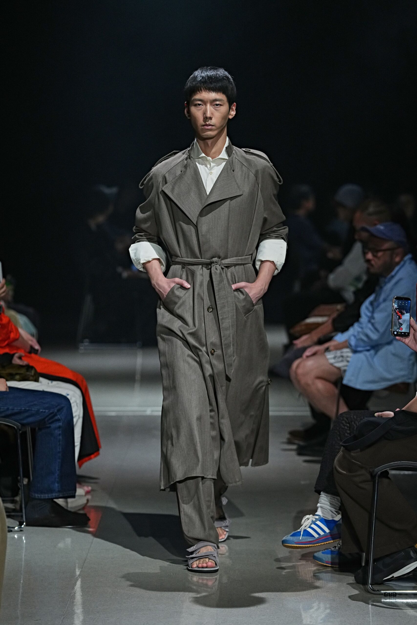 White cotton shirt, collar spread open and cuffs turned broad. Greige trench in dry suiting cloth, epaulettes drooping off the shoulders. Matching straight trousers breaking softly over the instep. Flat grey sandals with folded straps crossing the toes. For the slow walk back to a hotel after rain and room-service coffee.