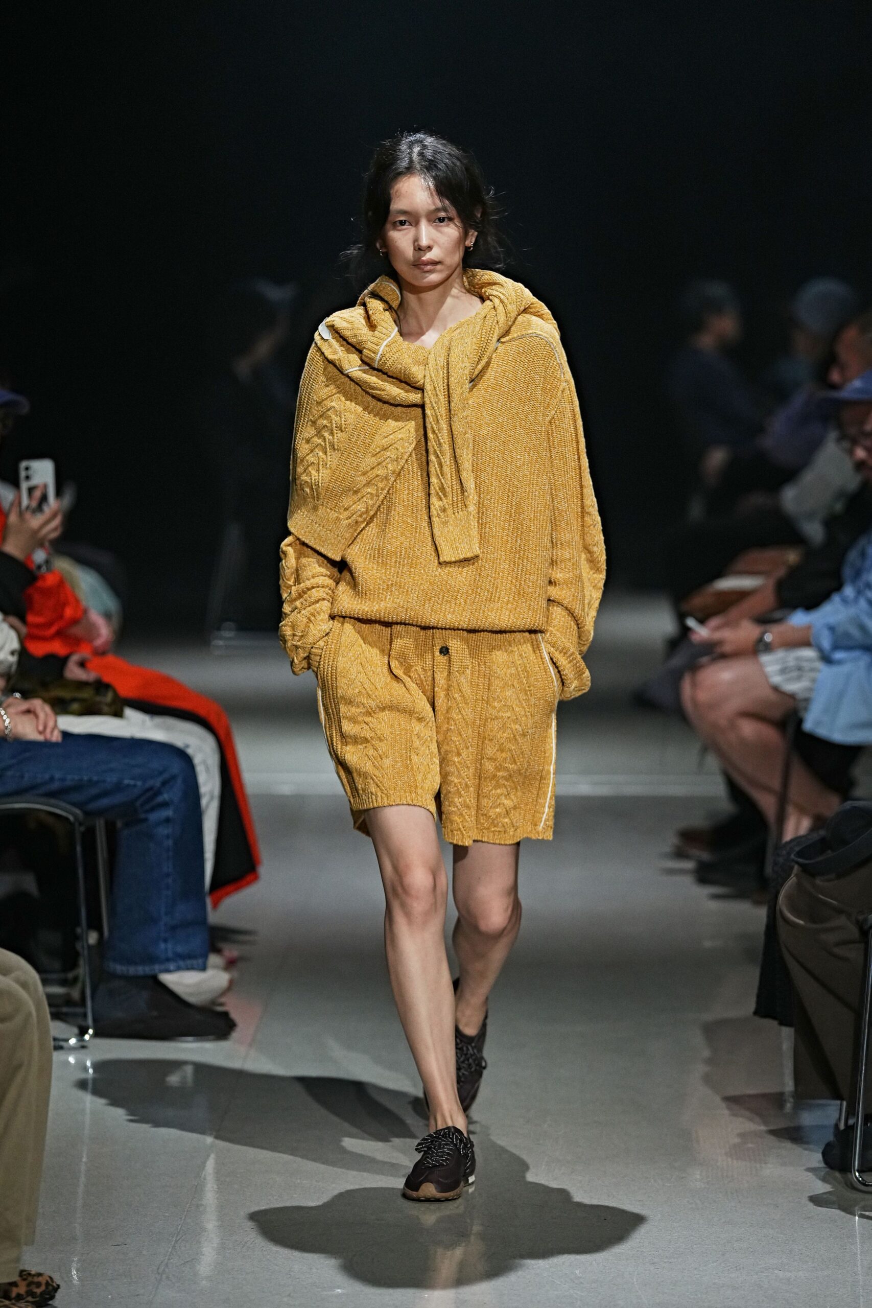 Ochre cable-knit sweater with a second knit looped thickly round the shoulders. Matching cable-knit shorts, one dark button peeking at the fly. Black woven shoes with chunky rubber soles. Cabin porch after rain, borrowing somebody else's cold-weather layers.