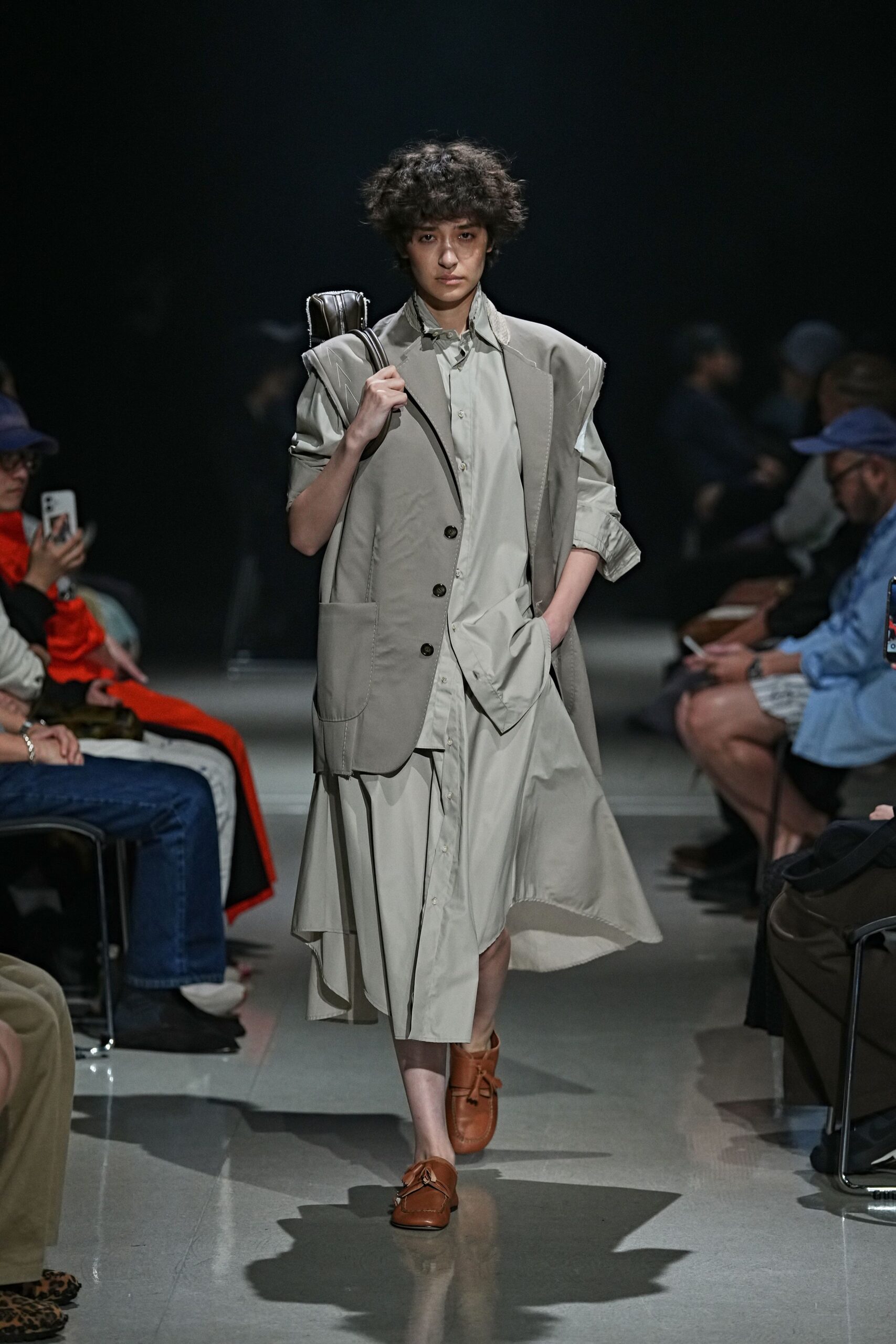 Slim black leather case slung upright against one shoulder. Greige tailored vest with cutaway armholes and oversized patch pockets. Pale poplin shirt dress, sleeves rolled unevenly, hem swinging below the knee. Cognac leather loafers with knotted apron laces. Walking to an early train with yesterday's shirt still half tucked.