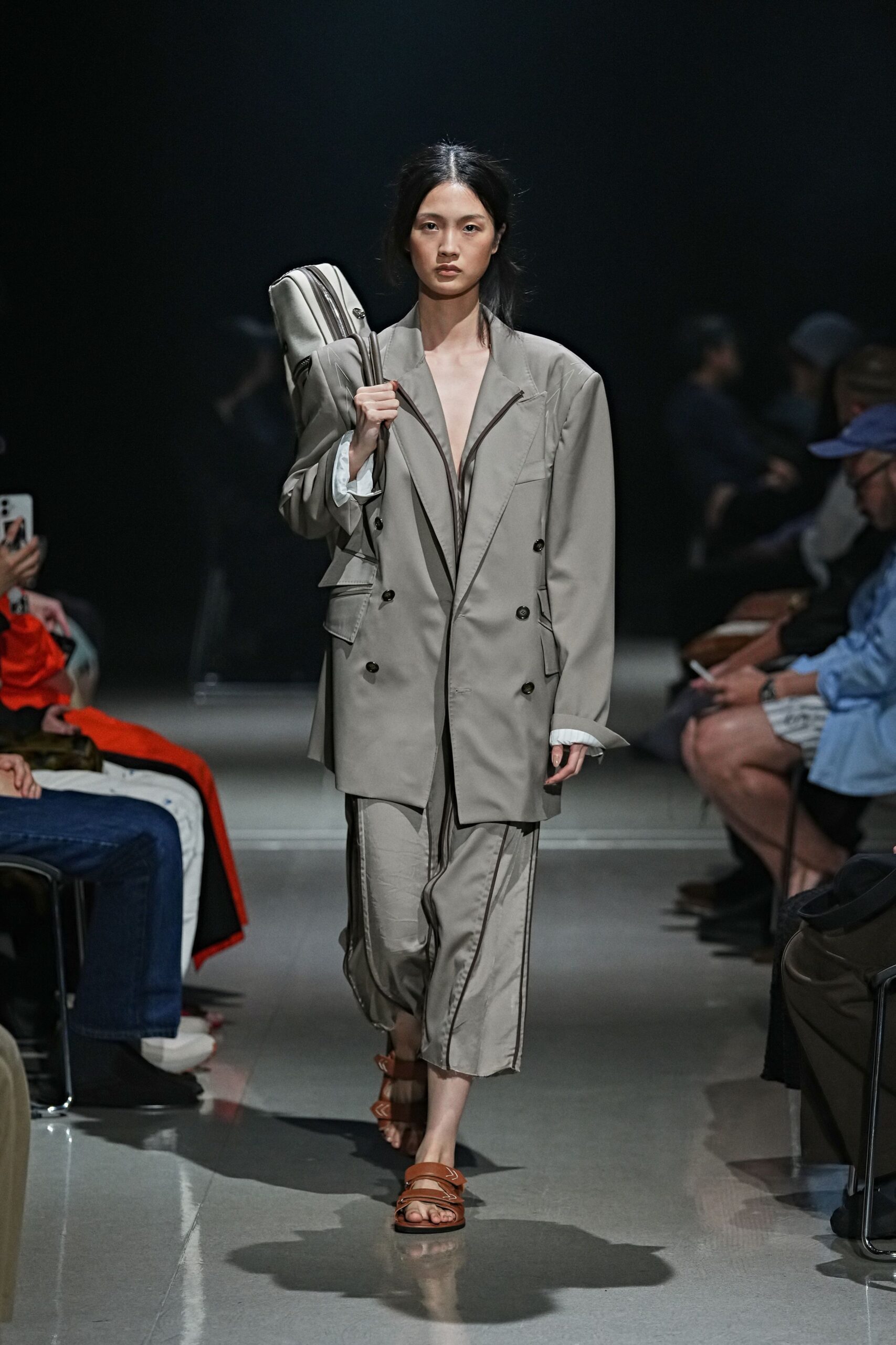 Oversized dove-grey blazer, double-breasted with a zip tracing the plunge. Bias-cut satin midi skirt with dark piping and a wavering hem. Tan leather slides with broad straps and white whipstitch marks. Long padded holdall slung on the shoulder. Walking to an early train with yesterday's suit jacket over a slip.