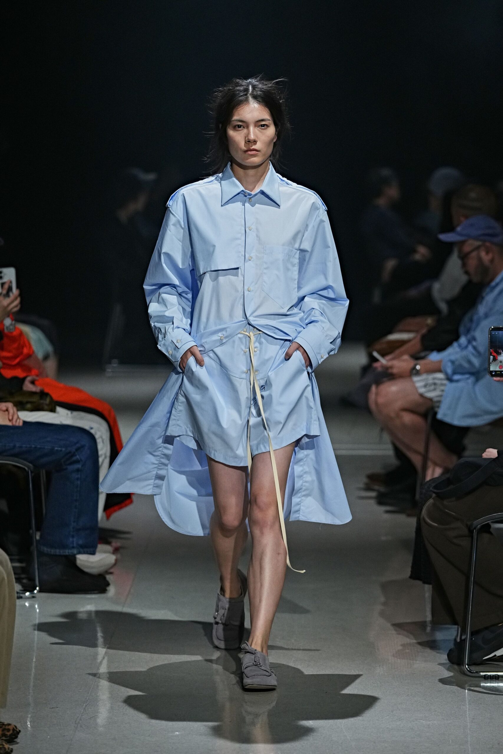 Powder-blue cotton shirting, dropped shoulder tabs and an uneven flap across the chest. Matching shirting shorts with a long cream drawcord hanging past the knee. Soft grey loafers with tassels collapsed flat over the vamp. Walking across an empty office floor before the lights fully come on.