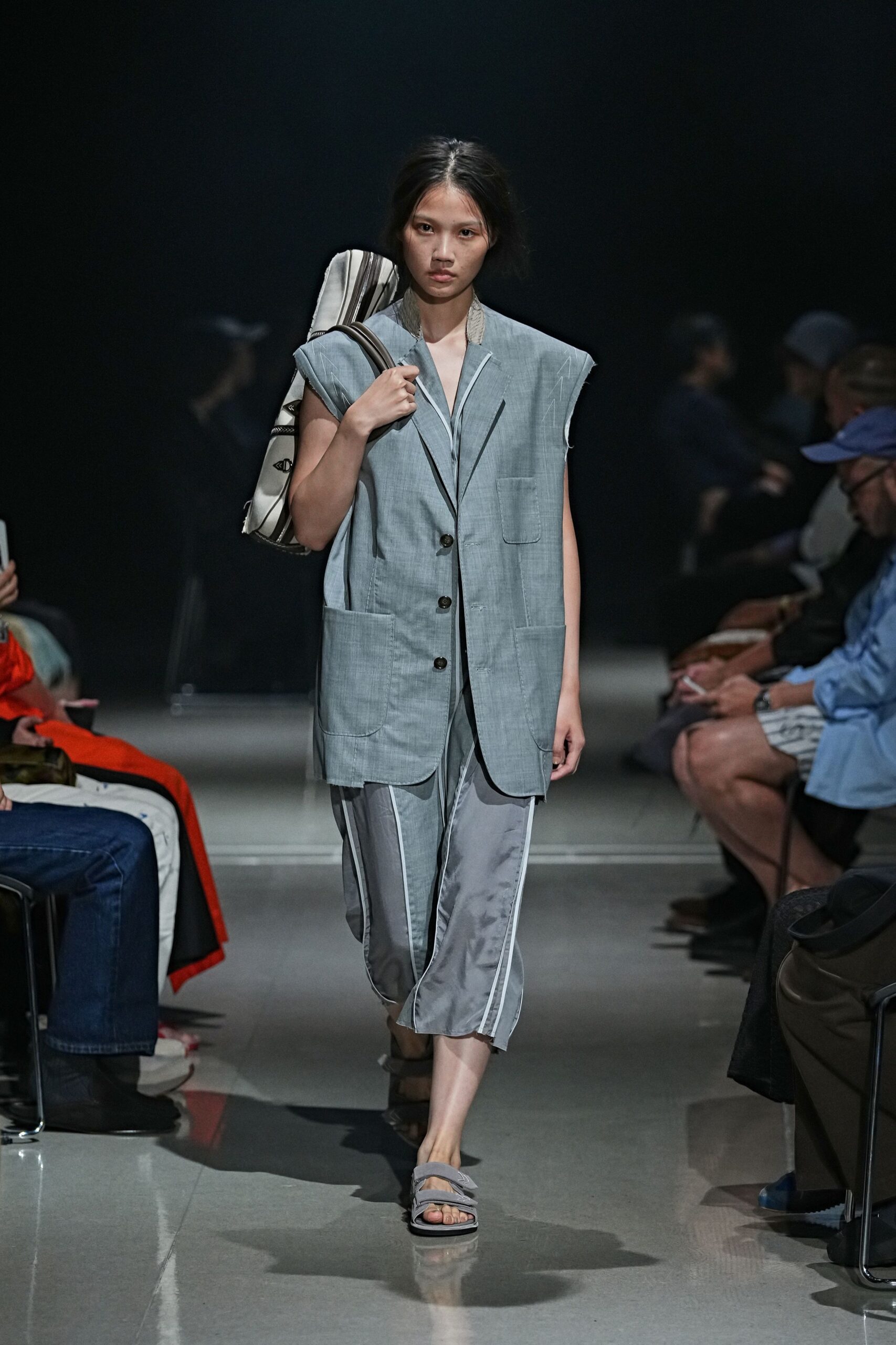 Pale blue chambray vest, raw armholes and oversized patch pockets. Silvery cropped trousers with white taped side seams and a liquid drape. Cream roll bag with black piping tucked under one arm. Grey padded slide sandals, double straps crossing low. For the walk from a station locker to rented rooms by the sea.