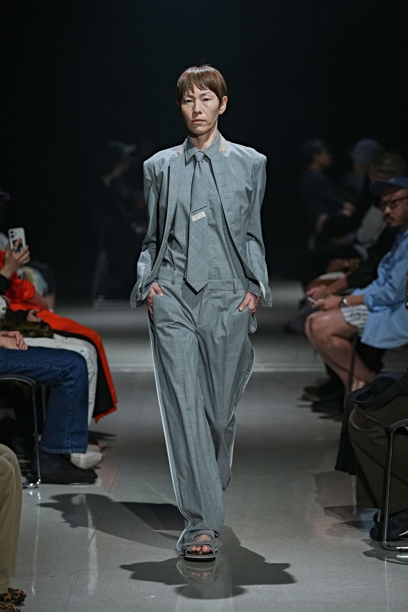 Pale grey suiting shirt with a long pointed collar and matching tie. Sleeveless open jacket in the same cloth, raw pale facings at the shoulders. Loose pleated trousers with white piping running the outer leg. Flat thong sandals with wrapped ankle ties. Early train to a design fair, carrying last night's creases with you.