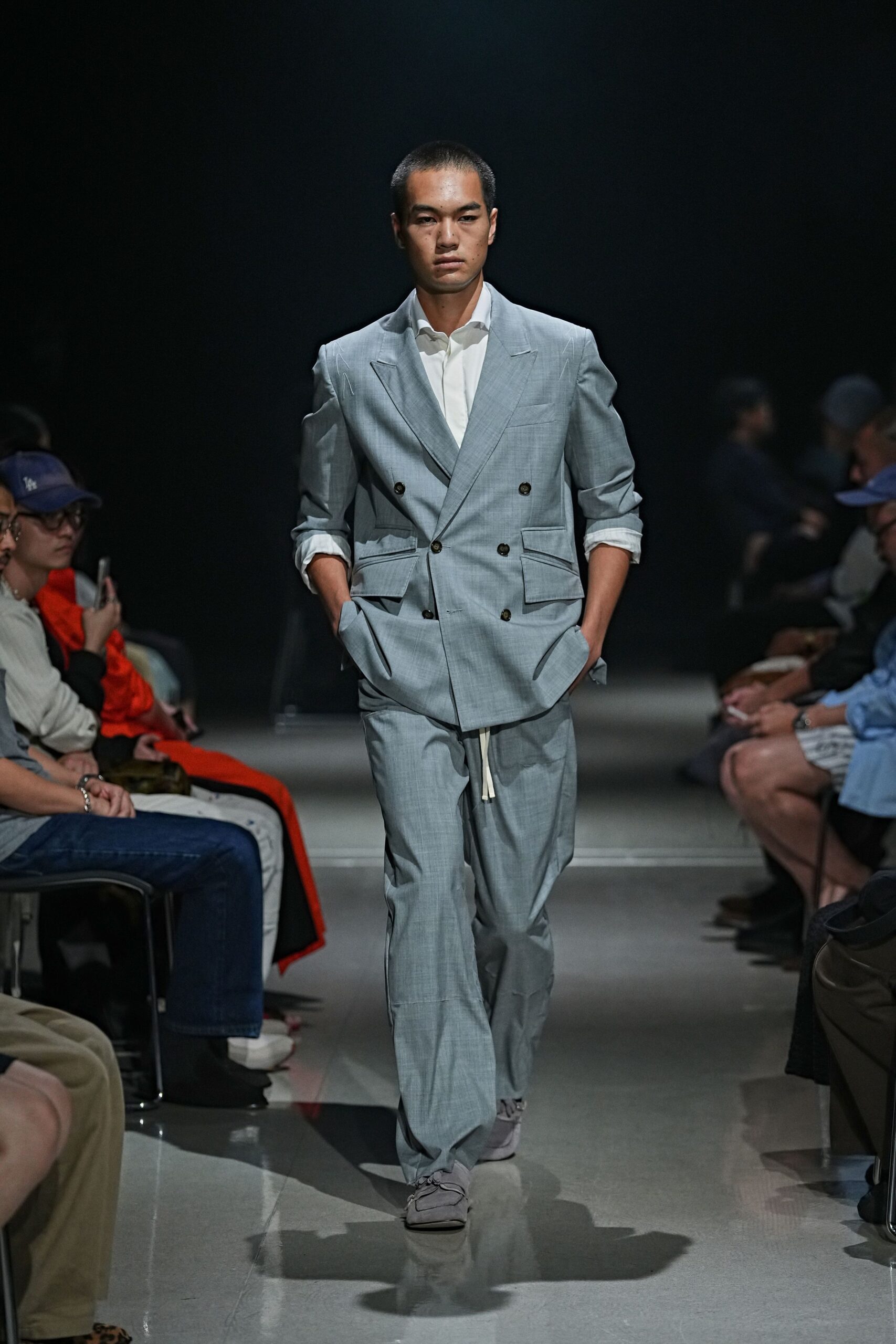 White shirt with the collar spread open and sleeves casually rolled. Pale blue double-breasted blazer, dark buttons and slanted flap pockets. Loose matching trousers with a cream drawstring hanging below the jacket. Soft grey lace-up shoes with squared moc-toe seams. For the train ride after a summer wedding, jacket still on.