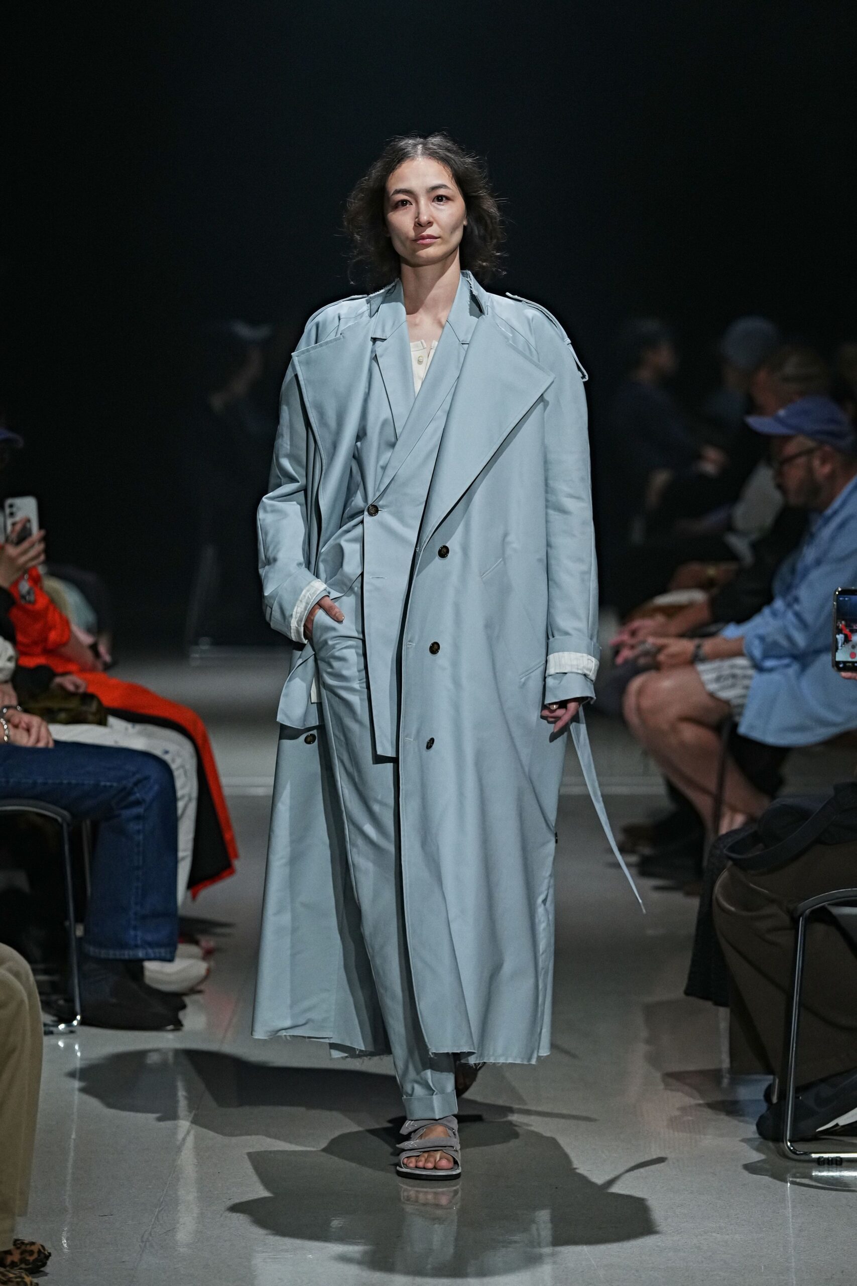 Powder-blue trench coat with oversized storm flaps and a loose belt end. Pale cream henley peeking through, small buttons at the throat. Matching blue trousers breaking in a soft cuff over the foot. Grey crossover sandals with padded straps. For an early train when the platform still smells of rain.