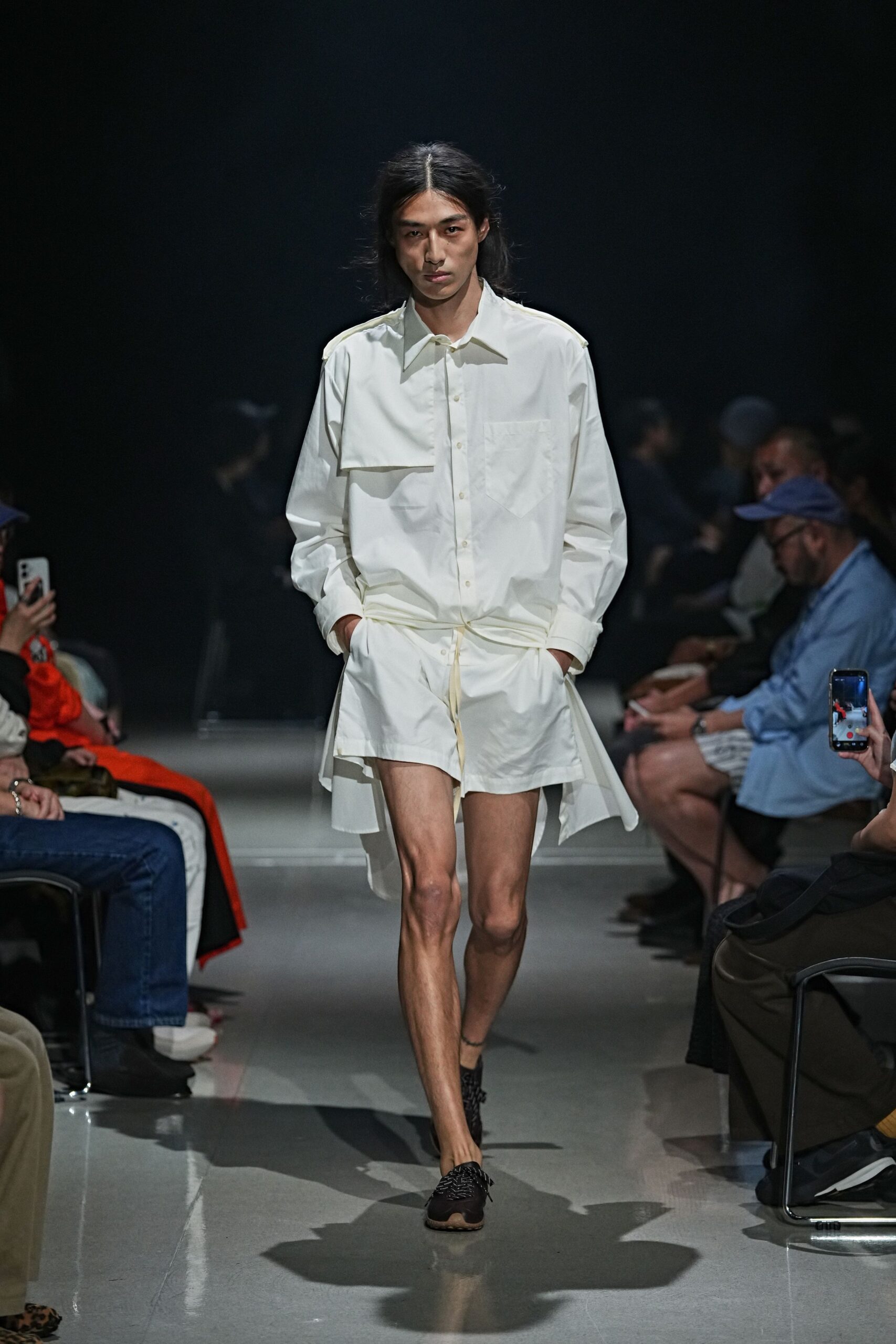 White cotton shirt with a floating bib panel and skewed chest pocket. Matching white shorts cinched with a thin drawstring at the waist. Black woven lace-up flats with pale rubber peeking under the toe. For crossing a hotel courtyard before anyone else is awake.