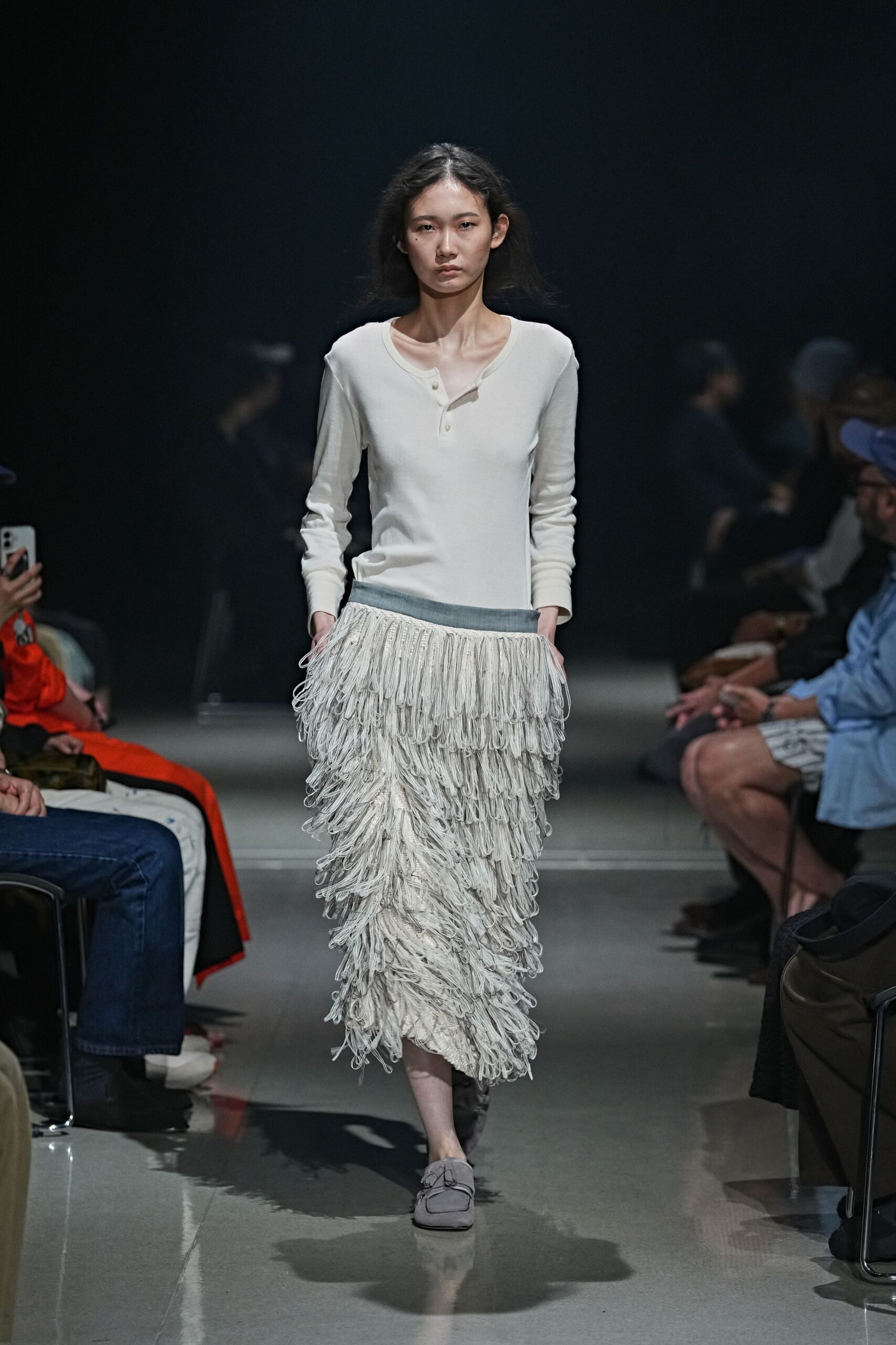 Ivory fine-rib henley, the placket tugged slightly off center. Long skirt banded in grey, covered in dense looped fringe tiers. Soft dove suede slippers with gathered ties at the vamp. Walking the hotel corridor before breakfast, carpet still marked by vacuum lines.