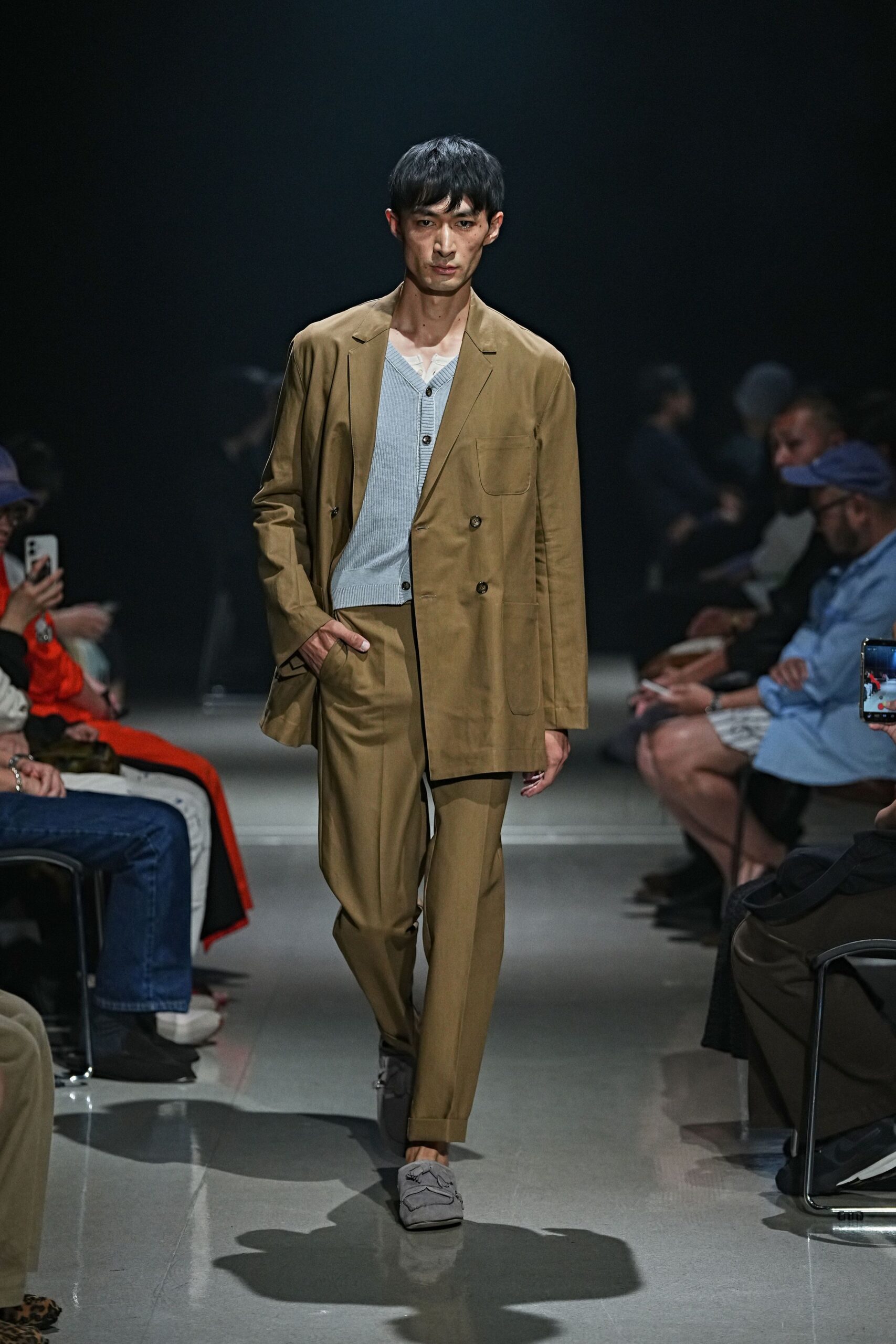 Khaki double-breasted blazer with rounded patch pockets and a softened drape. Pale blue ribbed cardigan, low V opening over a white undershirt edge. Matching khaki trousers with a pressed crease and turned-up hems. Dove grey suede loafers with rippling apron seams. For the first meeting after August when nobody wants a tie.