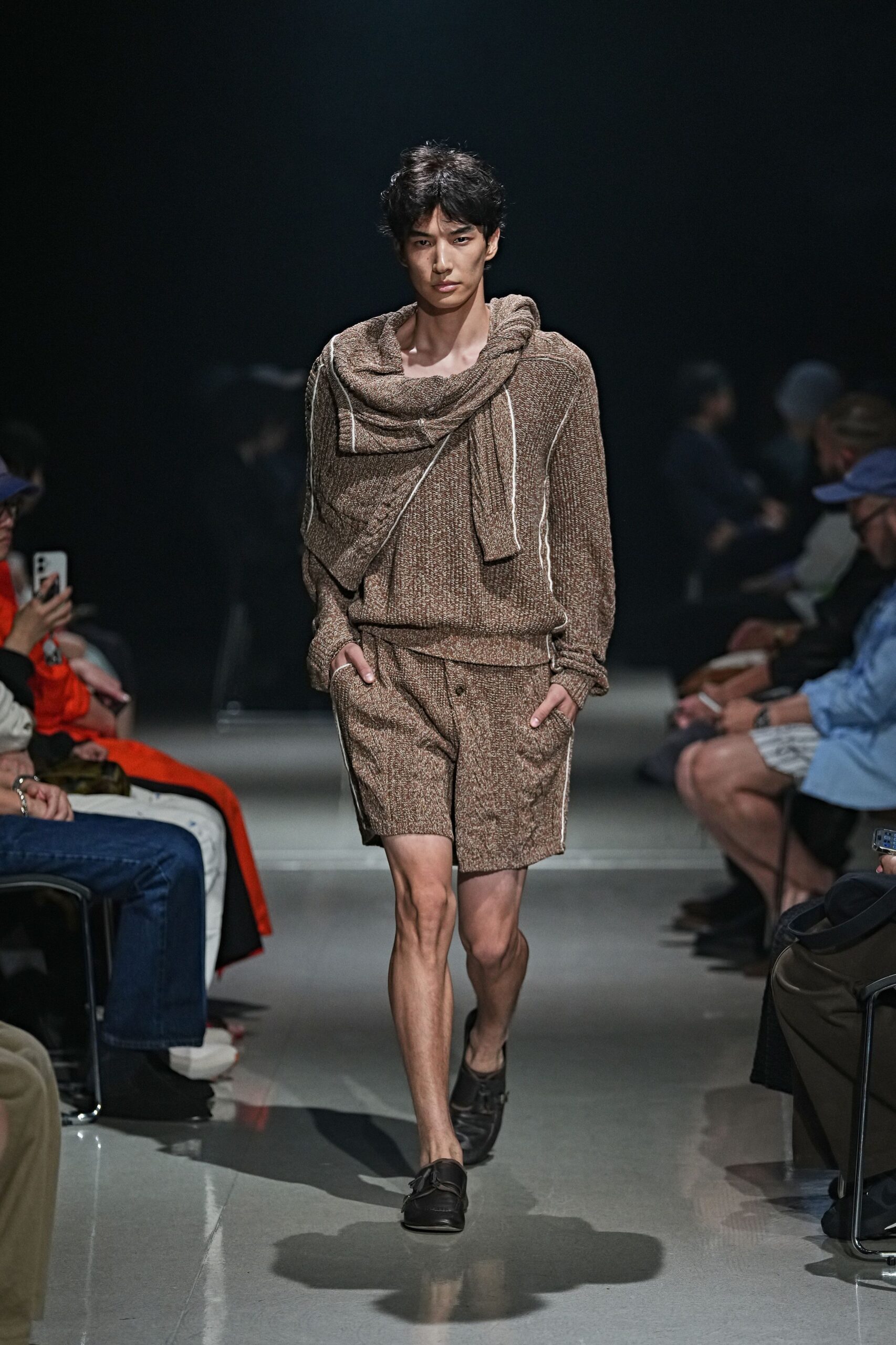 Brown marl knit sweater with white overlock lines running the raglan seams. Matching knit scarf looped wide across the shoulders, one end hanging long. Loose brown knit shorts with a low button tab at the fly. Black leather loafers with squared vamps. For buying figs at the market before the heat gets heavy.