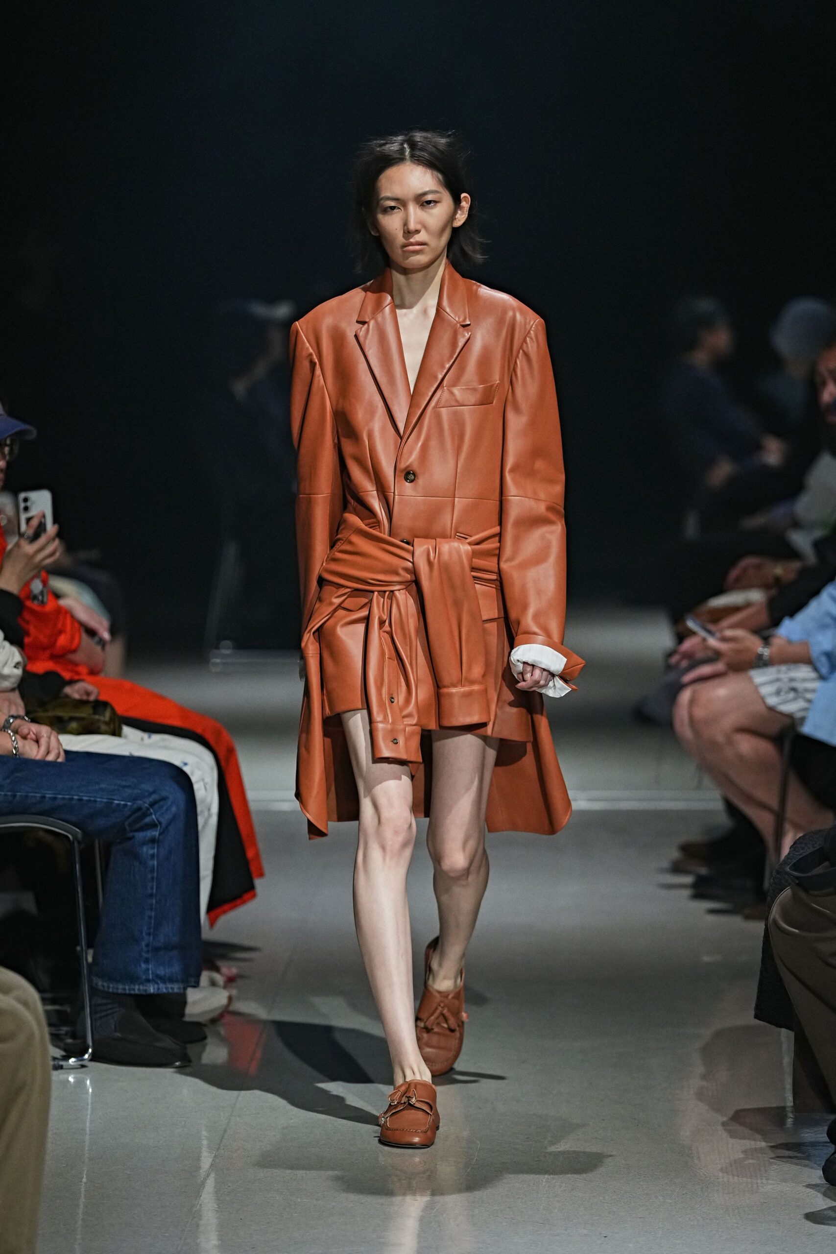 Cognac leather coatdress with broad shoulders and a shirt knotted hard at the waist. White cuff peeking out, wrinkled under the leather sleeve. Soft cognac loafers with tasseled apron seams. Whatever you pull on for a courthouse errand that turns into dinner.