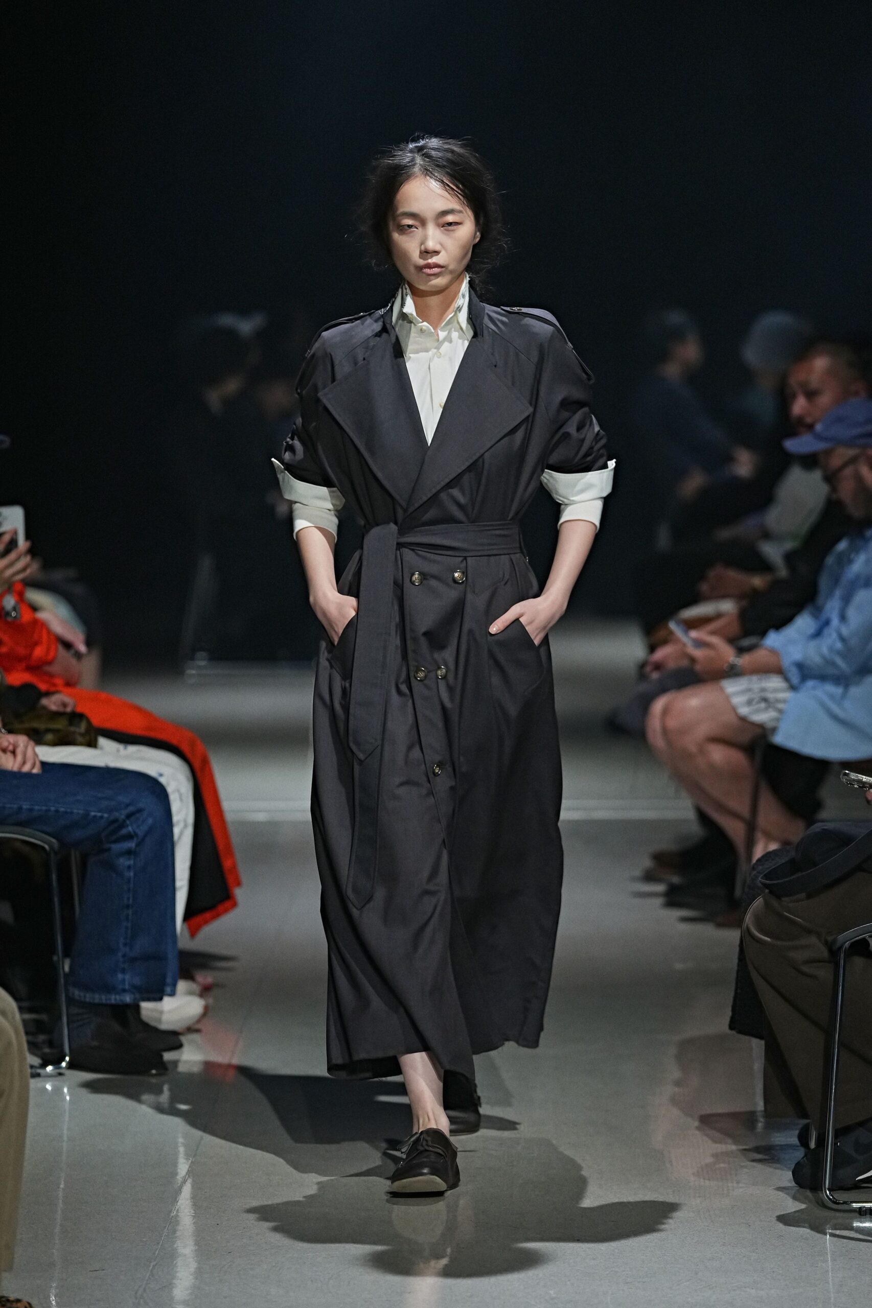 Ink twill trench dress with wide lapels and a long self-belt. White cotton shirt, cuffs turned back over the rolled coat sleeves. Black leather loafers with a soft collapsed vamp. For crossing the plaza early, before the office tower doors unlock.