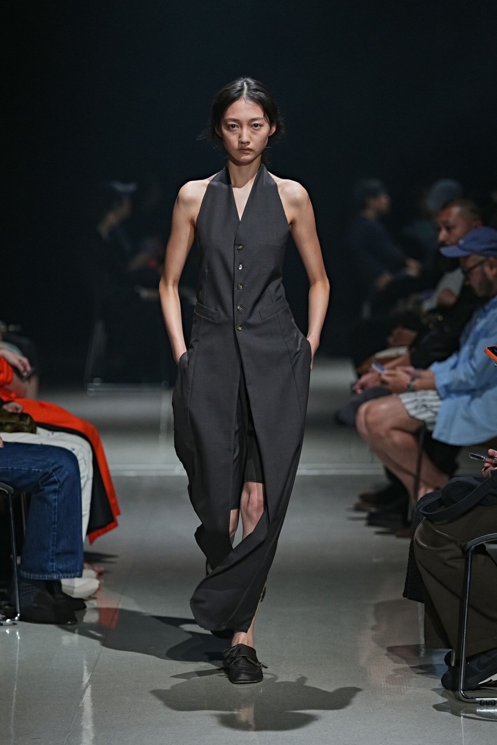 Charcoal halter jumpsuit with a deep V and mismatched brass buttons. Fluid wide legs split open below the pocket line. Black leather loafers, softly creased across the vamp. For crossing an empty hotel lobby before sunrise, keycard in hand.