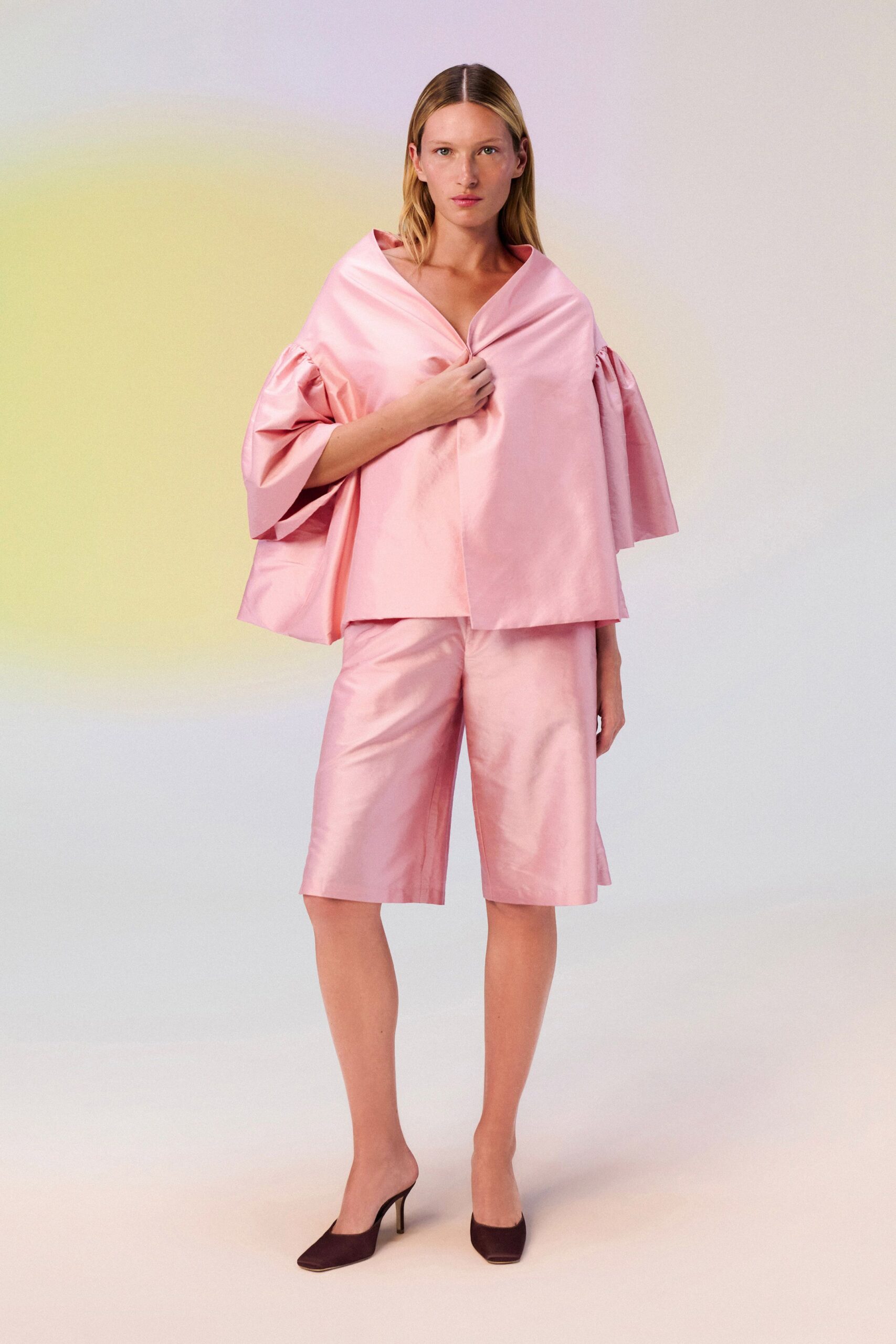 Blush duchesse satin cape top, pinched closed at the sternum. Matching satin bermudas cut wide and flat to the knee. Dark chocolate square-toe mules on slim stilettos. For crossing a hotel lobby at dawn with yesterday's bouquet still in hand.