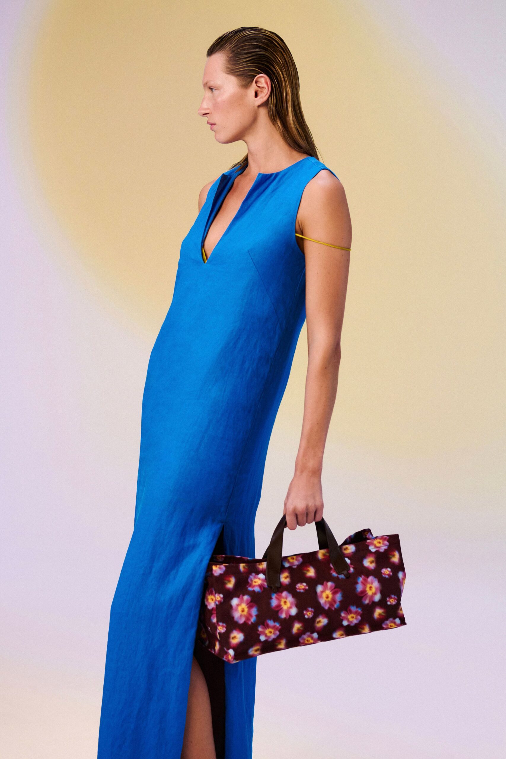 Cobalt column dress in crisp woven cloth, deep split V and side slit. Thin yellow cord looped once around the upper arm. Boxy tote in blurred burgundy floral print with dark leather handles. Walking back from a hotel pool bar with someone else's paperback.