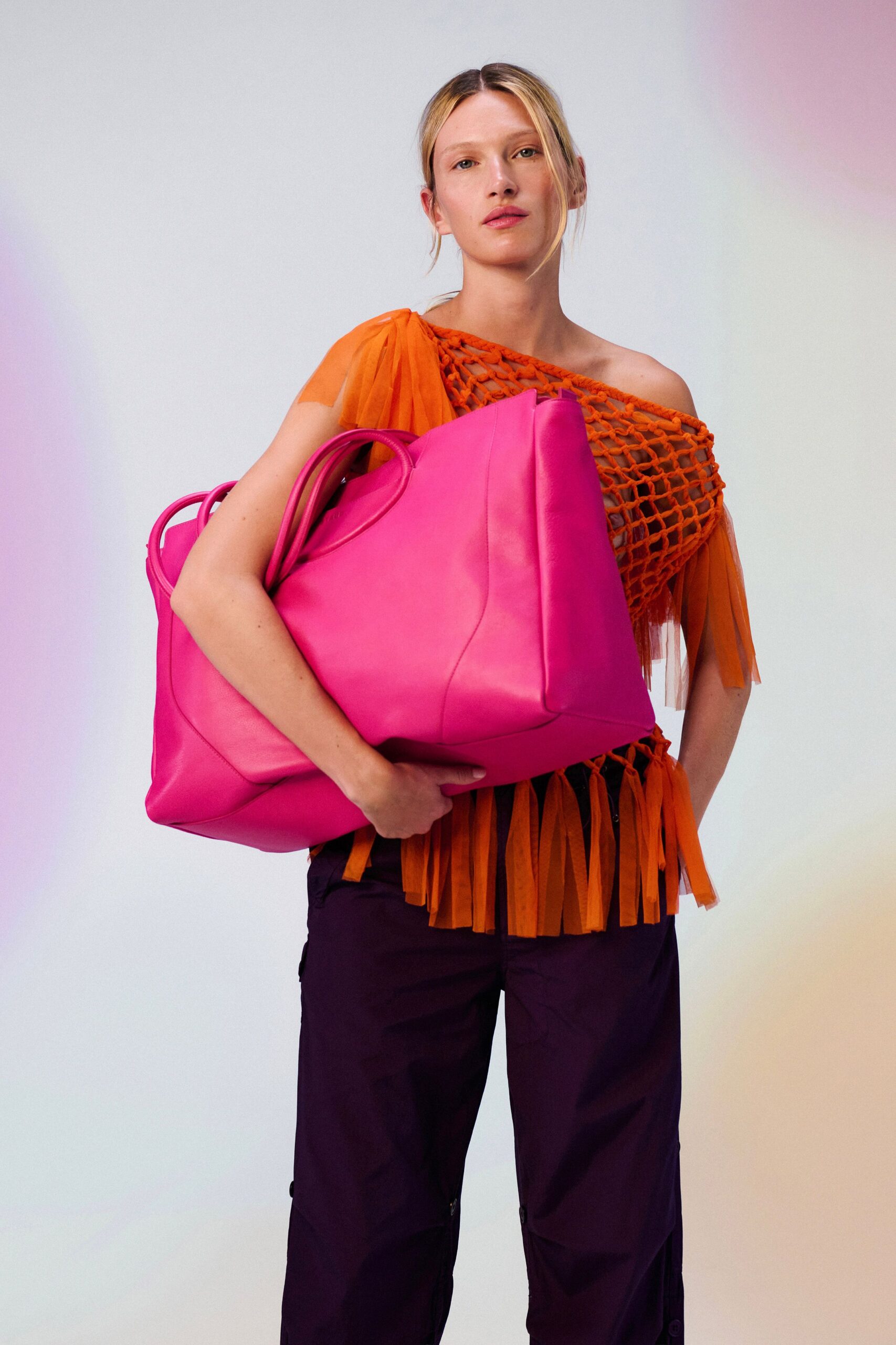 Orange open-net top slung off one shoulder, chiffon streamers fringing the hem. Oversized fuchsia leather tote with doubled handles and a softly collapsing side panel. Deep aubergine nylon cargo pants, zip fly peeking beneath the fringe. Walking back from the beach market with too much fruit in one arm.