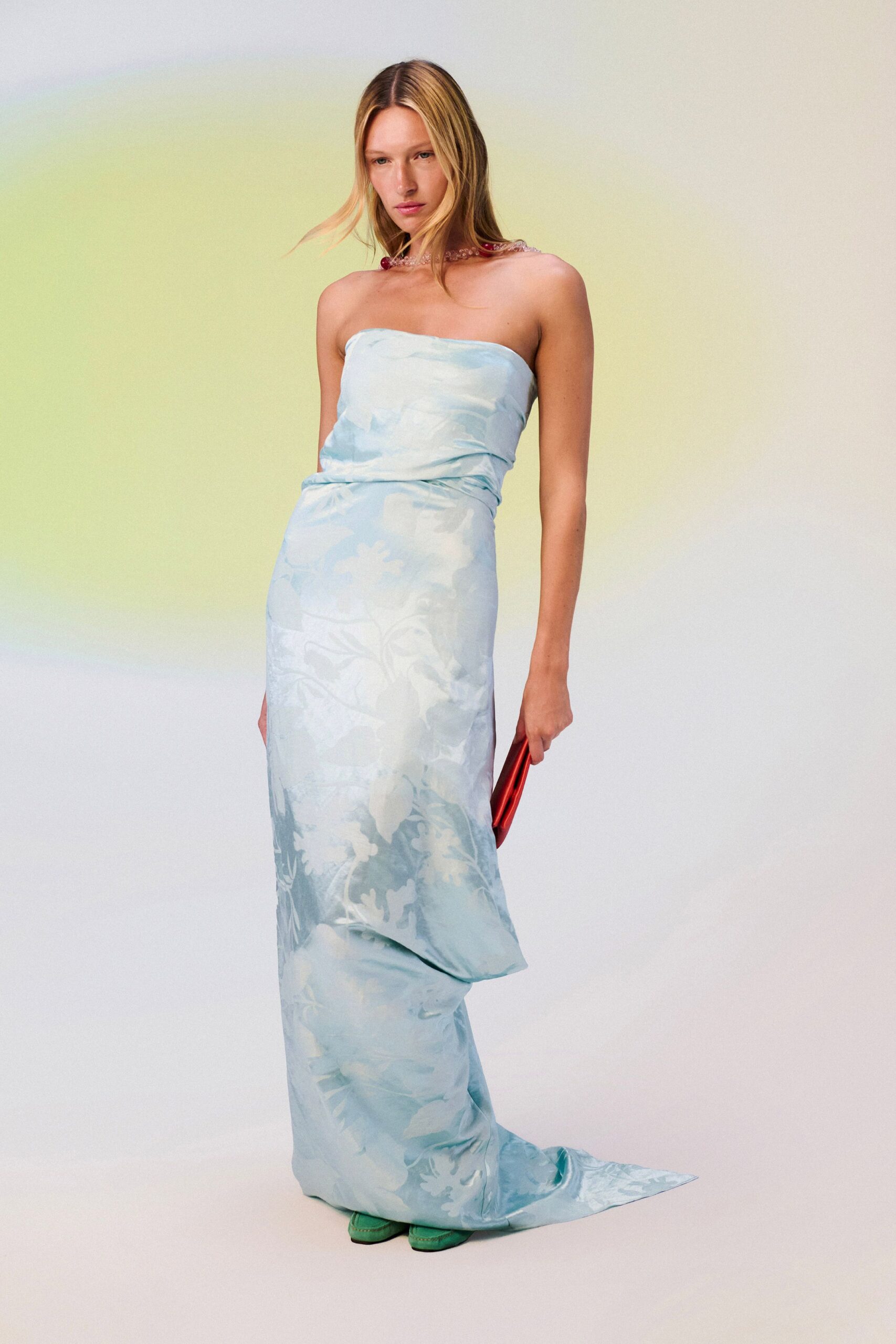 Pink beaded choker with a deeper red cluster at one shoulder. Strapless ice-blue satin column, tonal floral jacquard and a twisted waist. Metallic scarlet clutch tucked flat in one hand. Green satin shoes peeking under the pooled hem. Hotel corridor at sunrise, carrying your shoes back from the wedding.